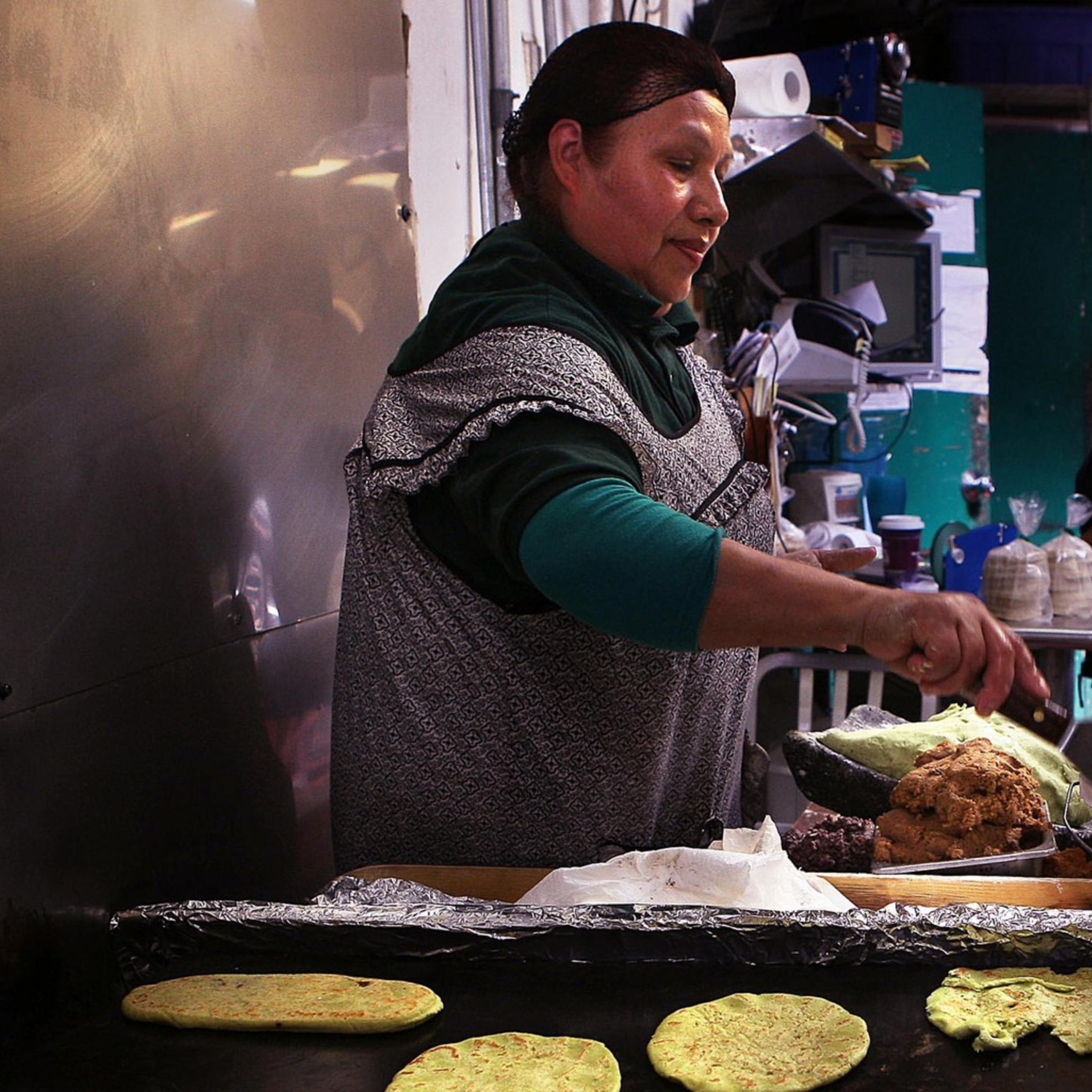 A woman in an apron cooks tortillas on a large griddle in a kitchen. Several people are in the background, and containers are nearby.