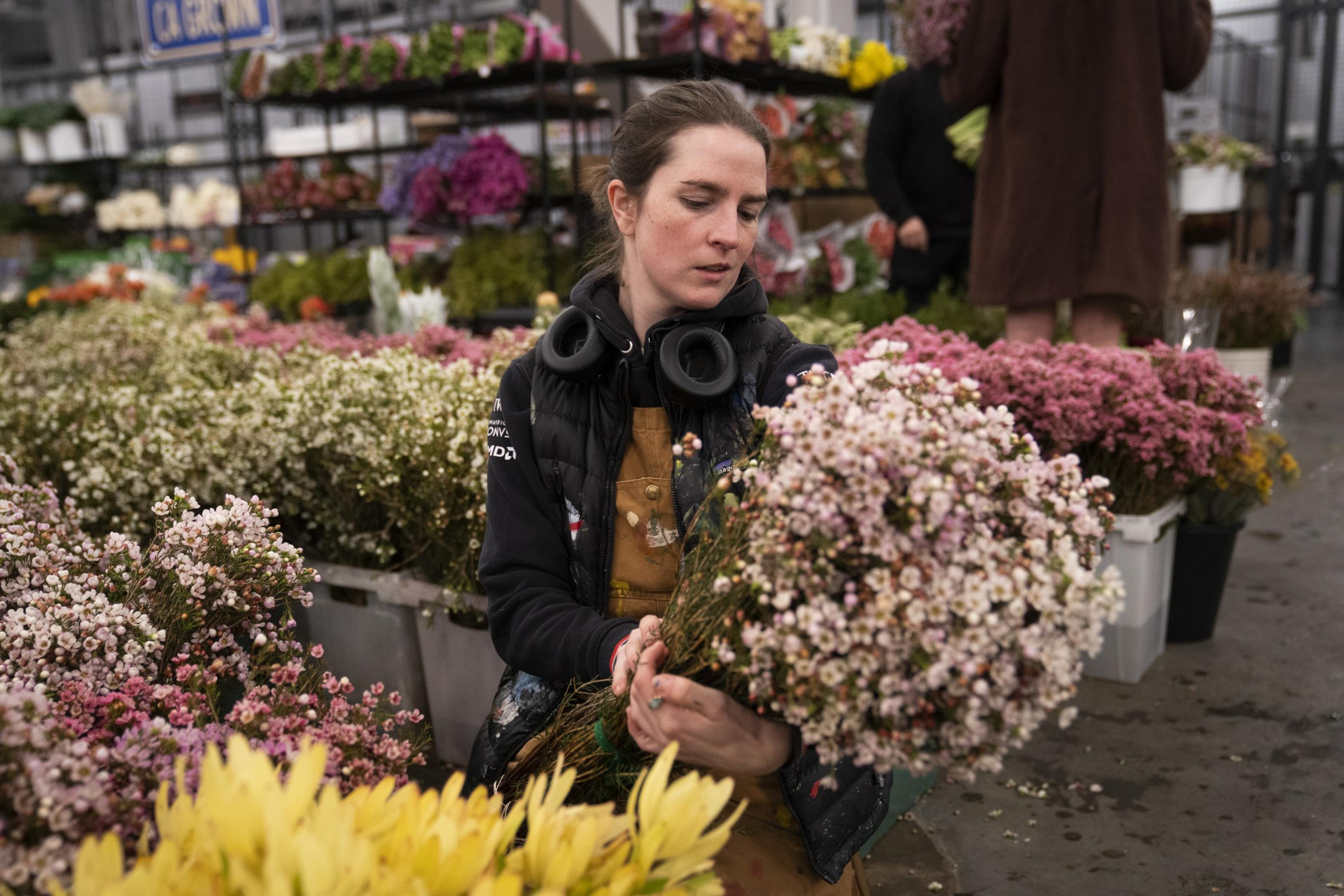 A person in overalls and a black jacket holds a bunch of white and pink flowers in a bustling market, surrounded by vibrant flower arrangements.