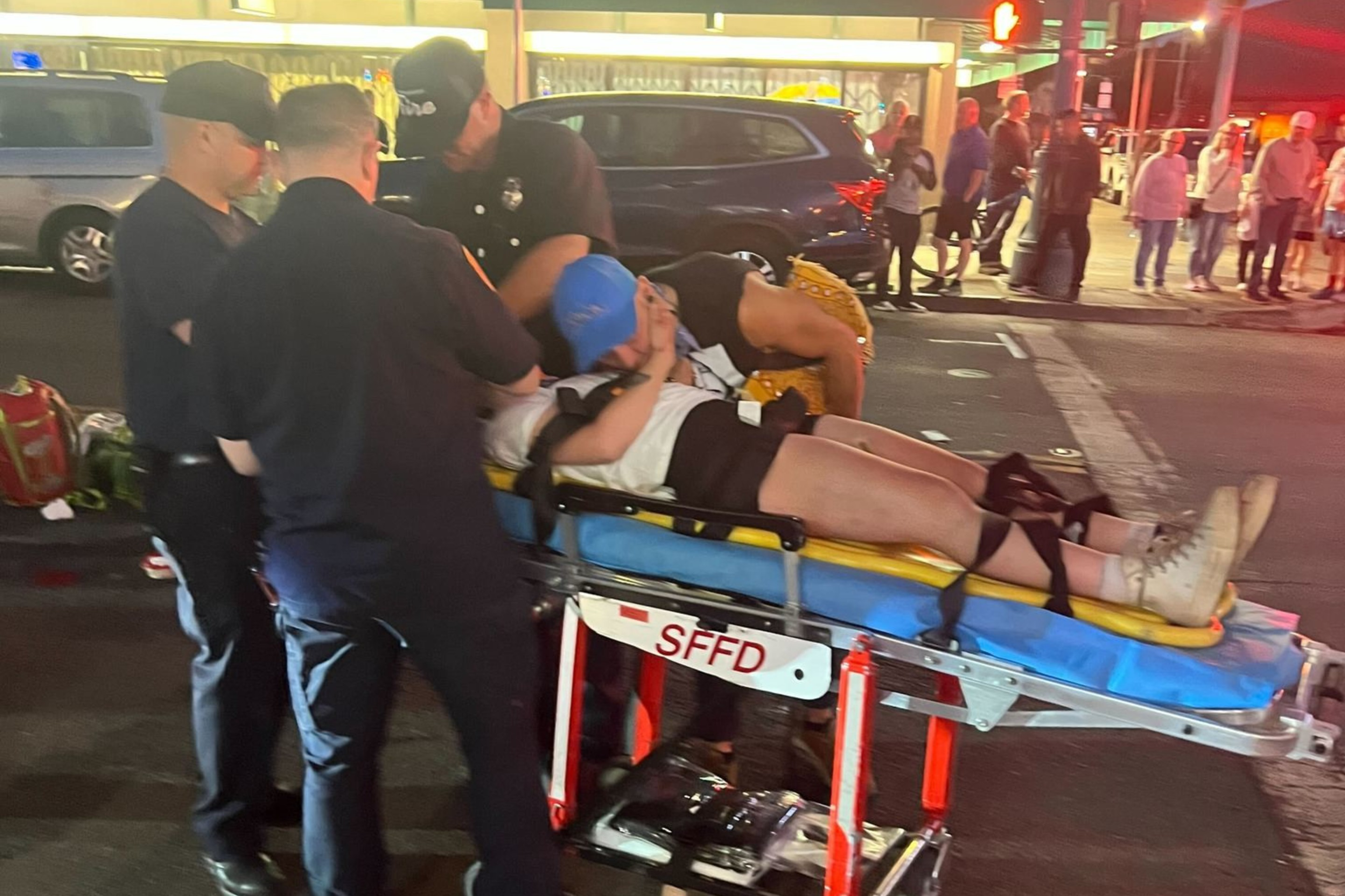 A person is lying on a stretcher attended by emergency responders in dark uniforms, on a street with bystanders nearby. The scene appears to be at night.