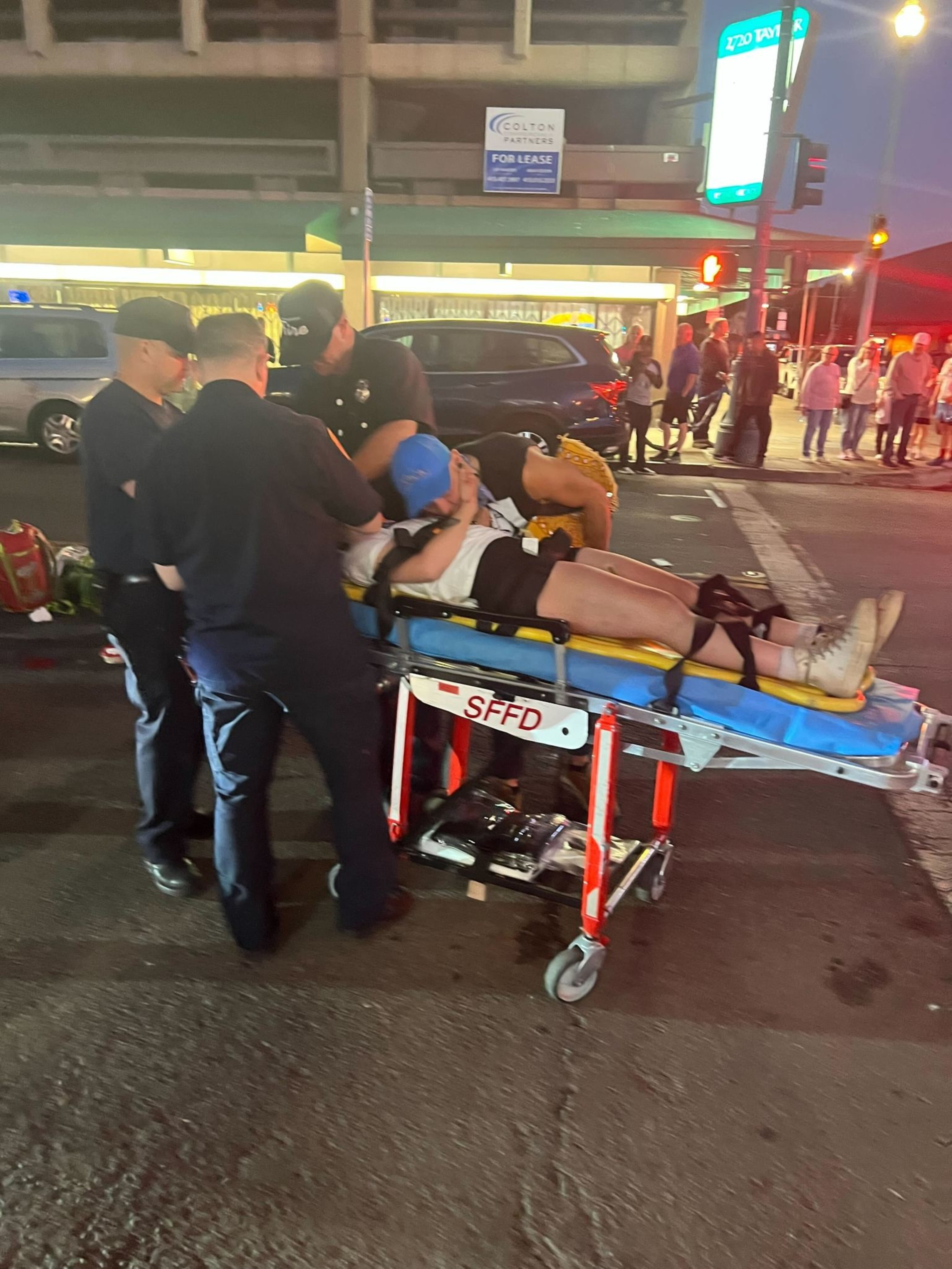 A person is lying on a stretcher attended by emergency responders in dark uniforms, on a street with bystanders nearby. The scene appears to be at night.