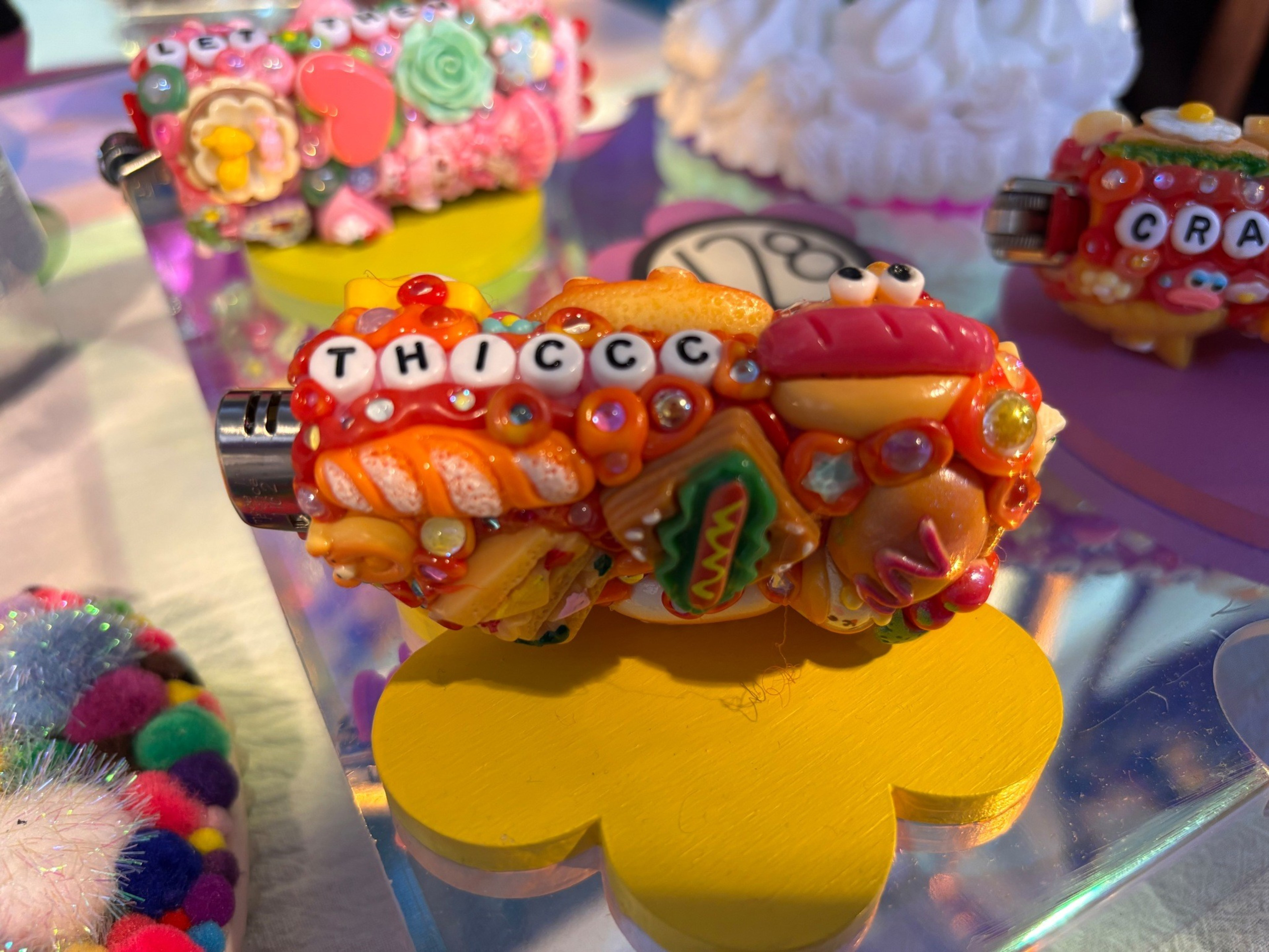A colorful decorated lighter with charms resembling food items and beads spelling “THICCC,” resting on a yellow flower-shaped stand.