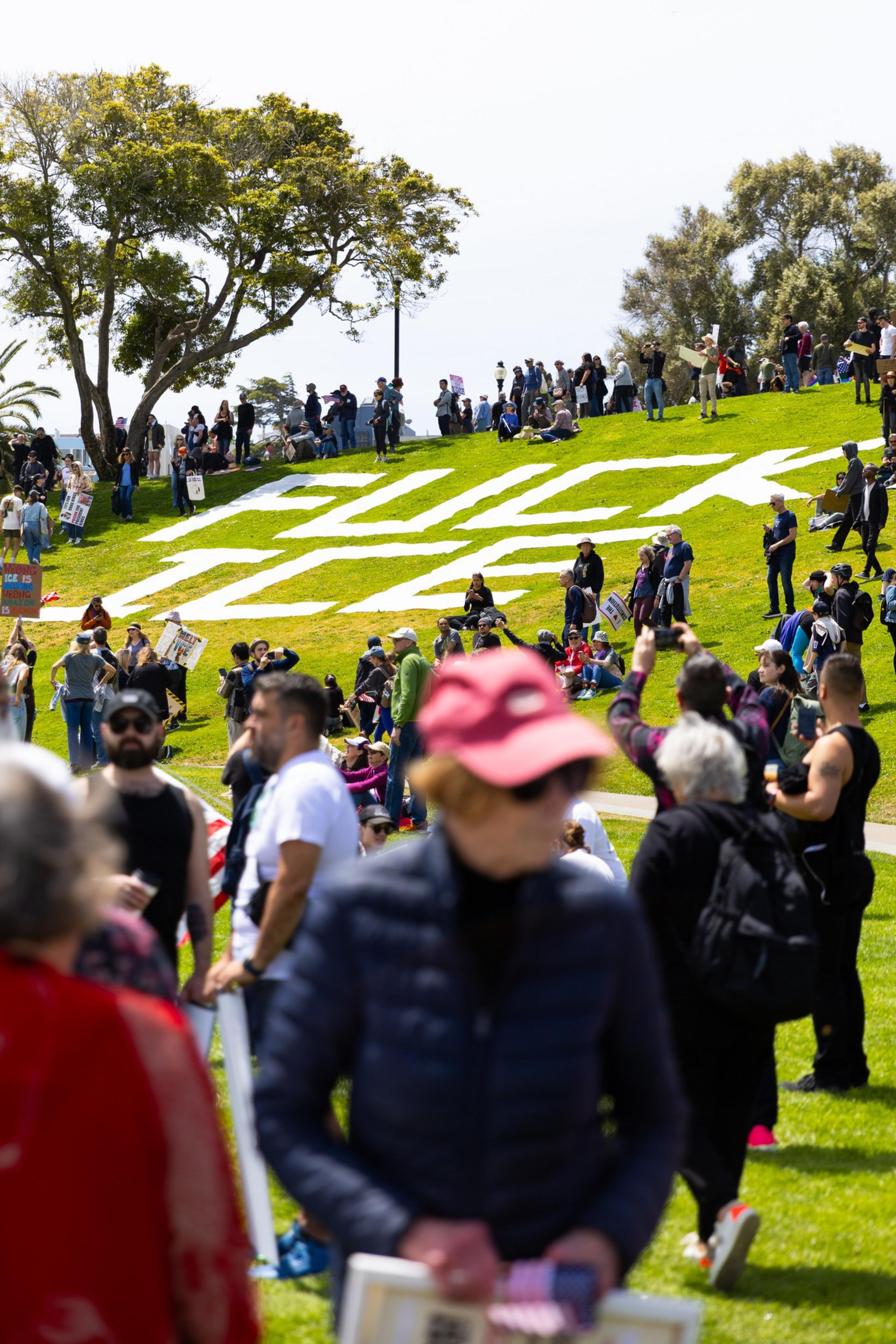 People gather on a sunny grassy hill with large white letters, surrounded by trees, some holding signs and taking photos.