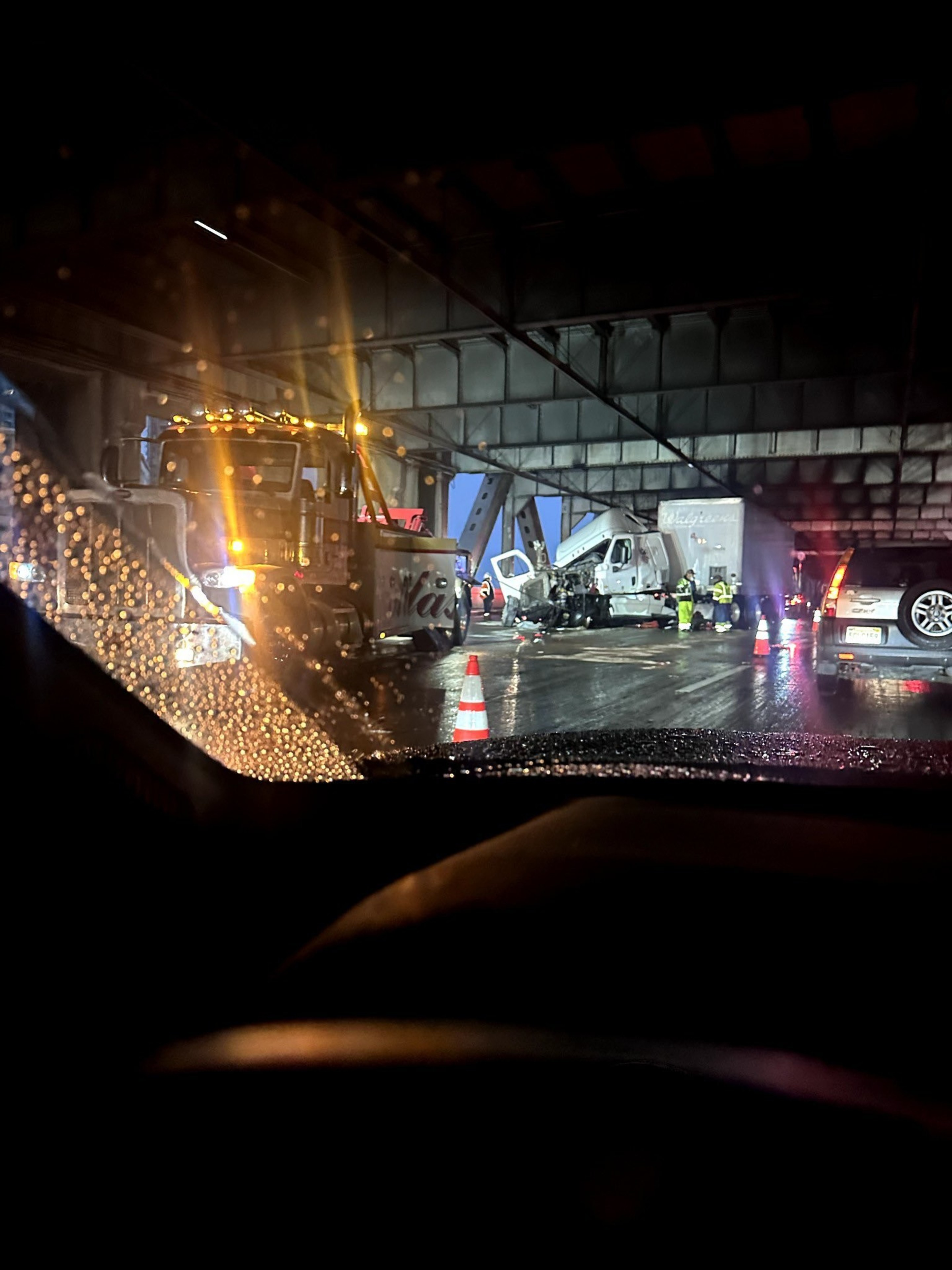 A night scene shows a truck accident under a bridge. Emergency workers are present, and traffic cones are placed around the area. It's raining, and road conditions are wet.