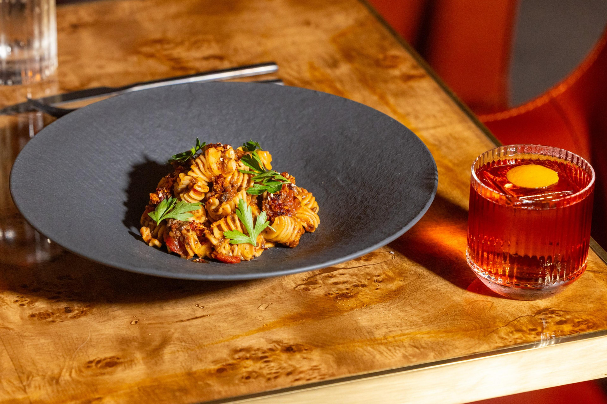 A black plate holds twisted pasta with tomato sauce and fresh parsley, placed on a wooden table next to a glass of amber cocktail with a lemon peel.