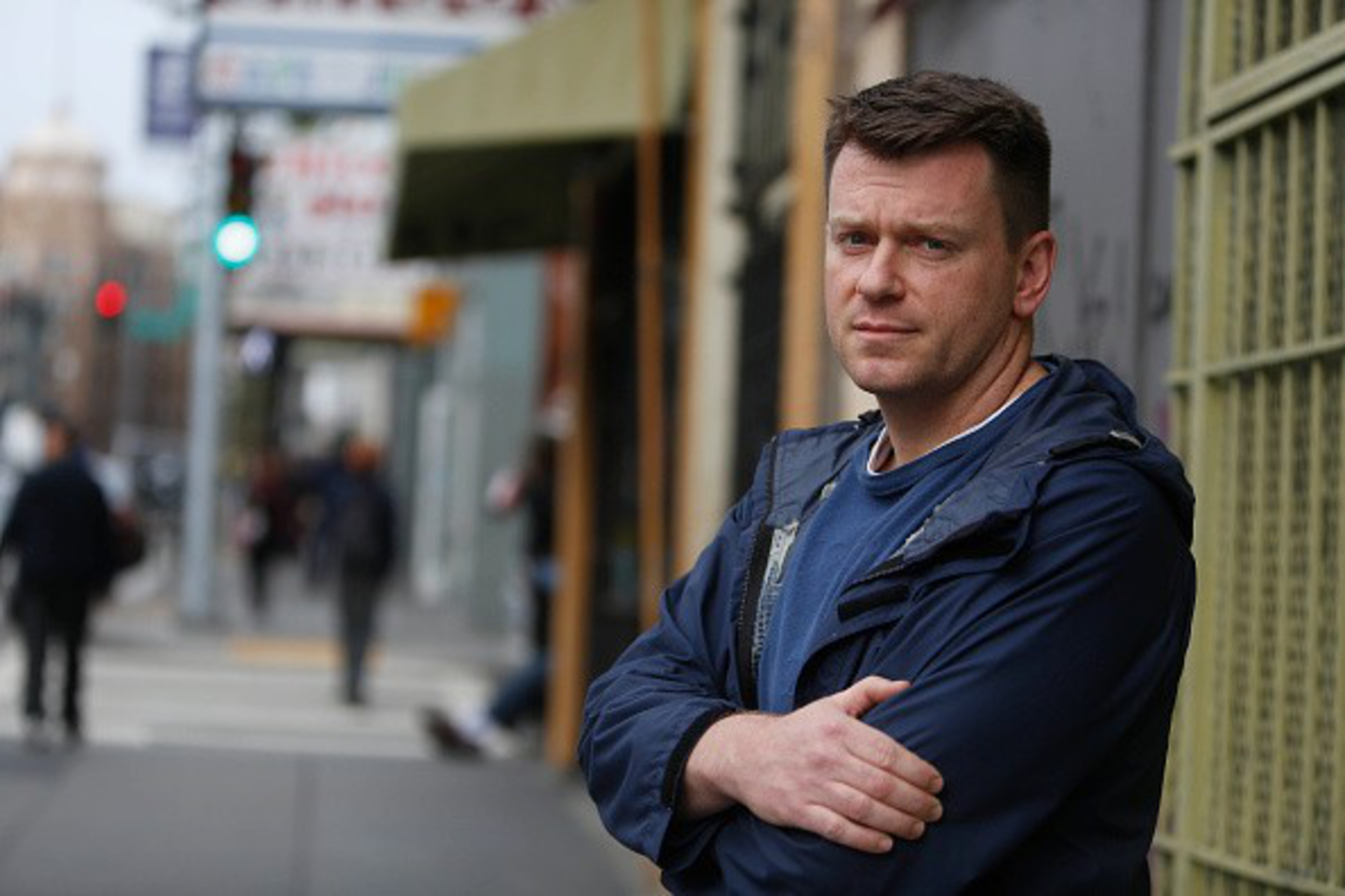 A man in a blue jacket stands with crossed arms on a city sidewalk, looking at the camera with a neutral expression.
