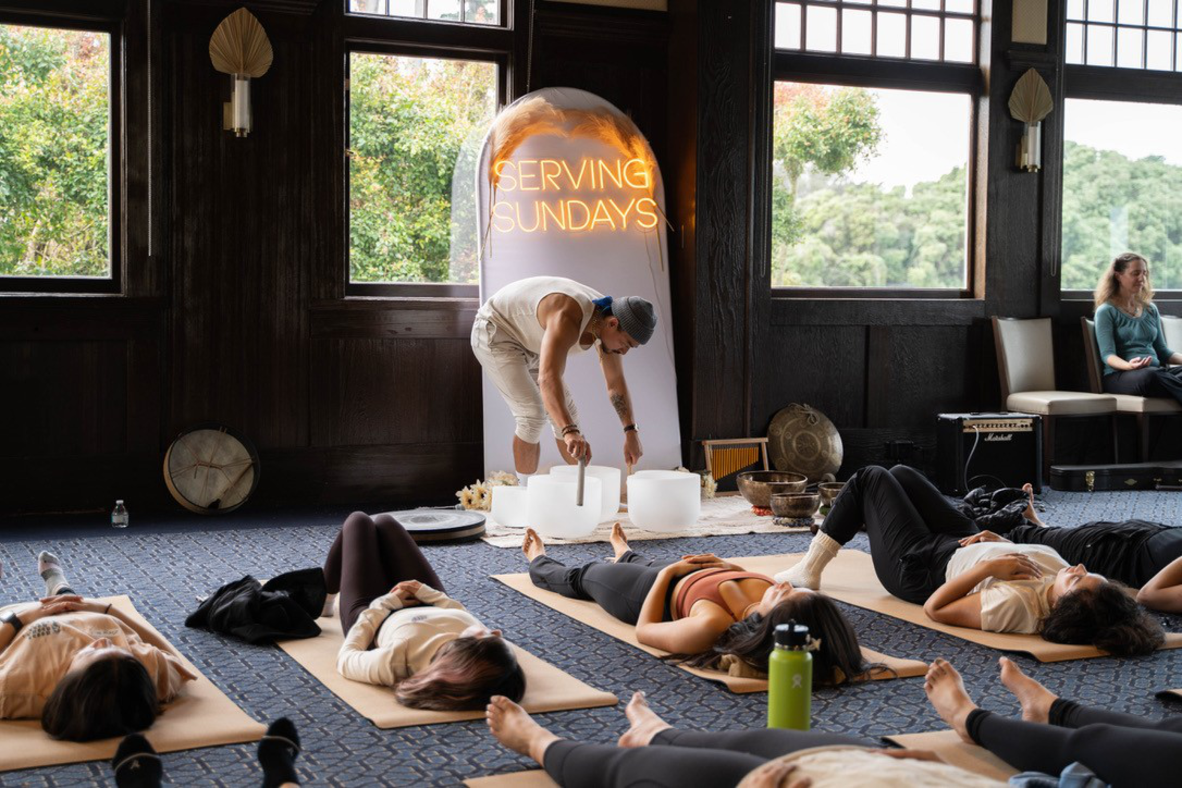 A group of people lie on yoga mats in a dimly lit room. A person is playing crystal singing bowls under a sign that reads &quot;SERVING SUNDAYS.&quot;