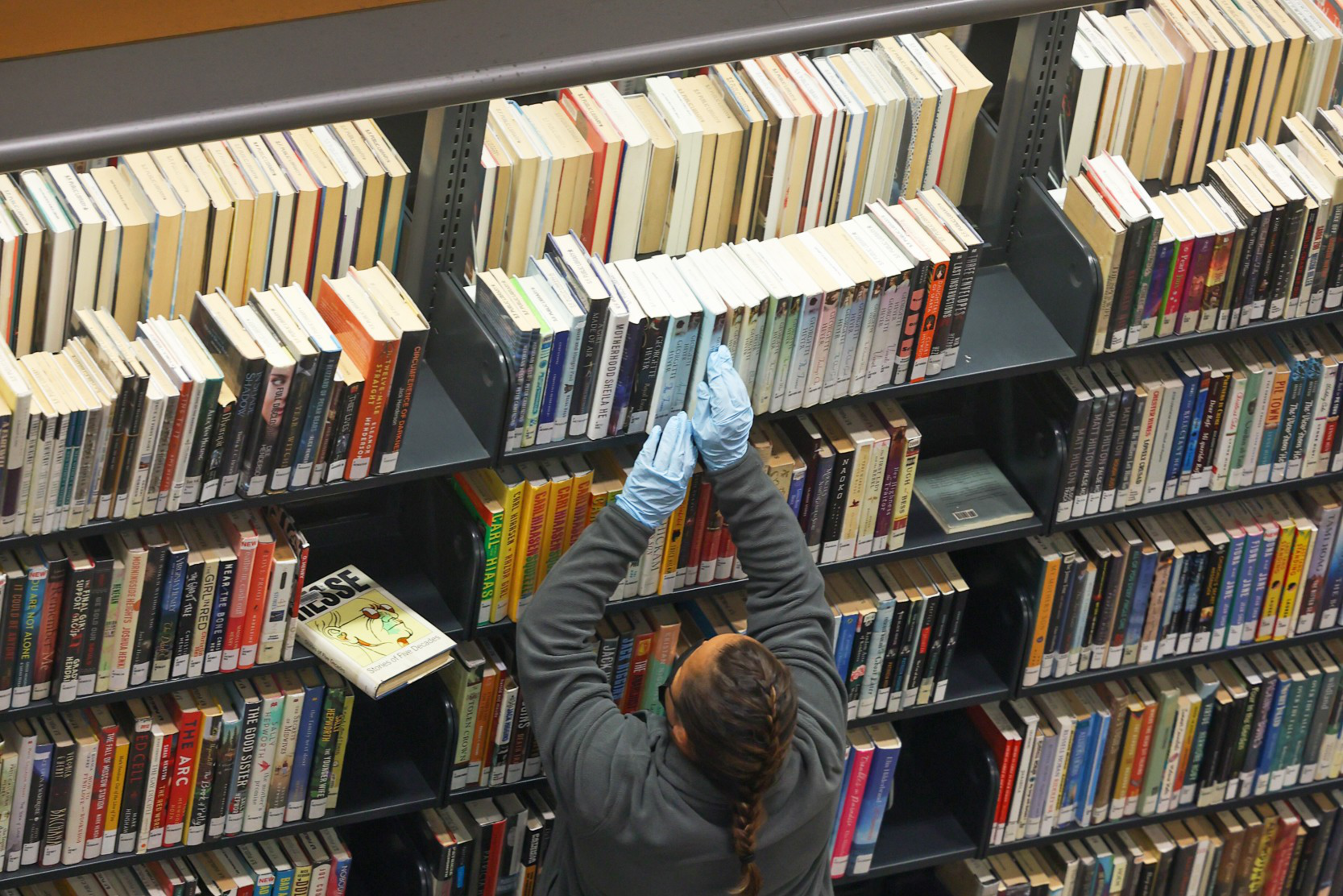 A person wearing blue gloves and a gray jacket is reaching for books on a high library shelf filled with various books stacked closely together.