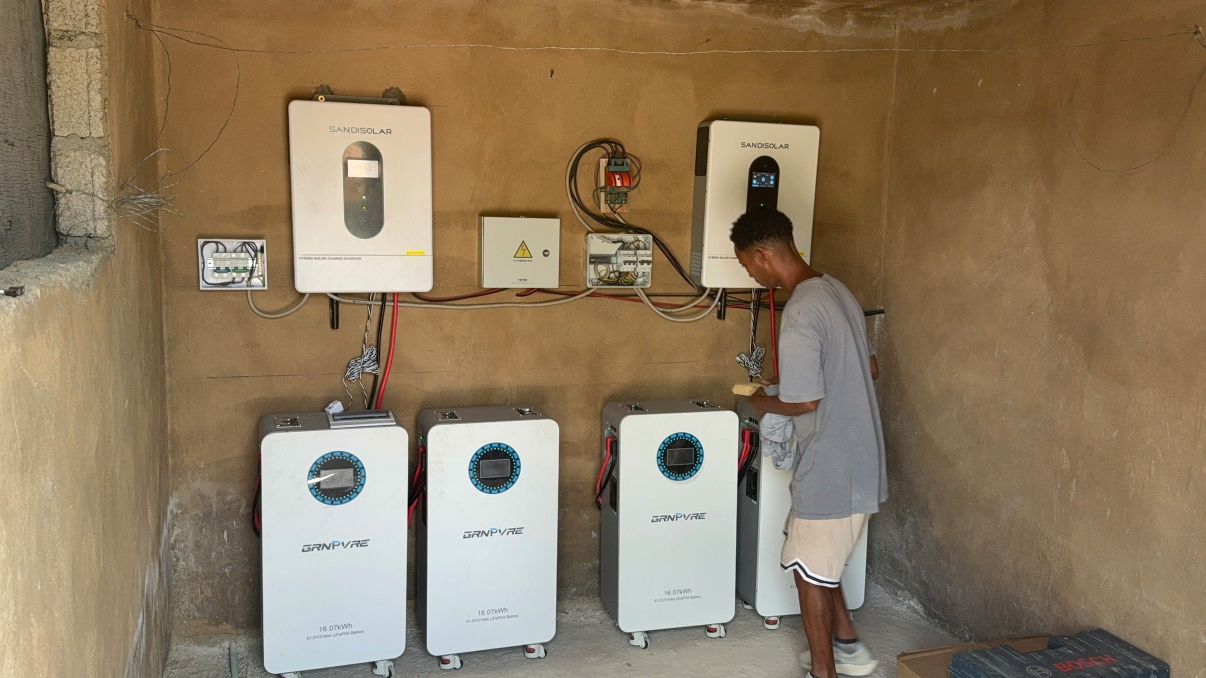 A man inspects four white battery units and mounted solar inverters labeled “SANDISOLAR” and “GRANPVIRE” inside a small room with bare walls.