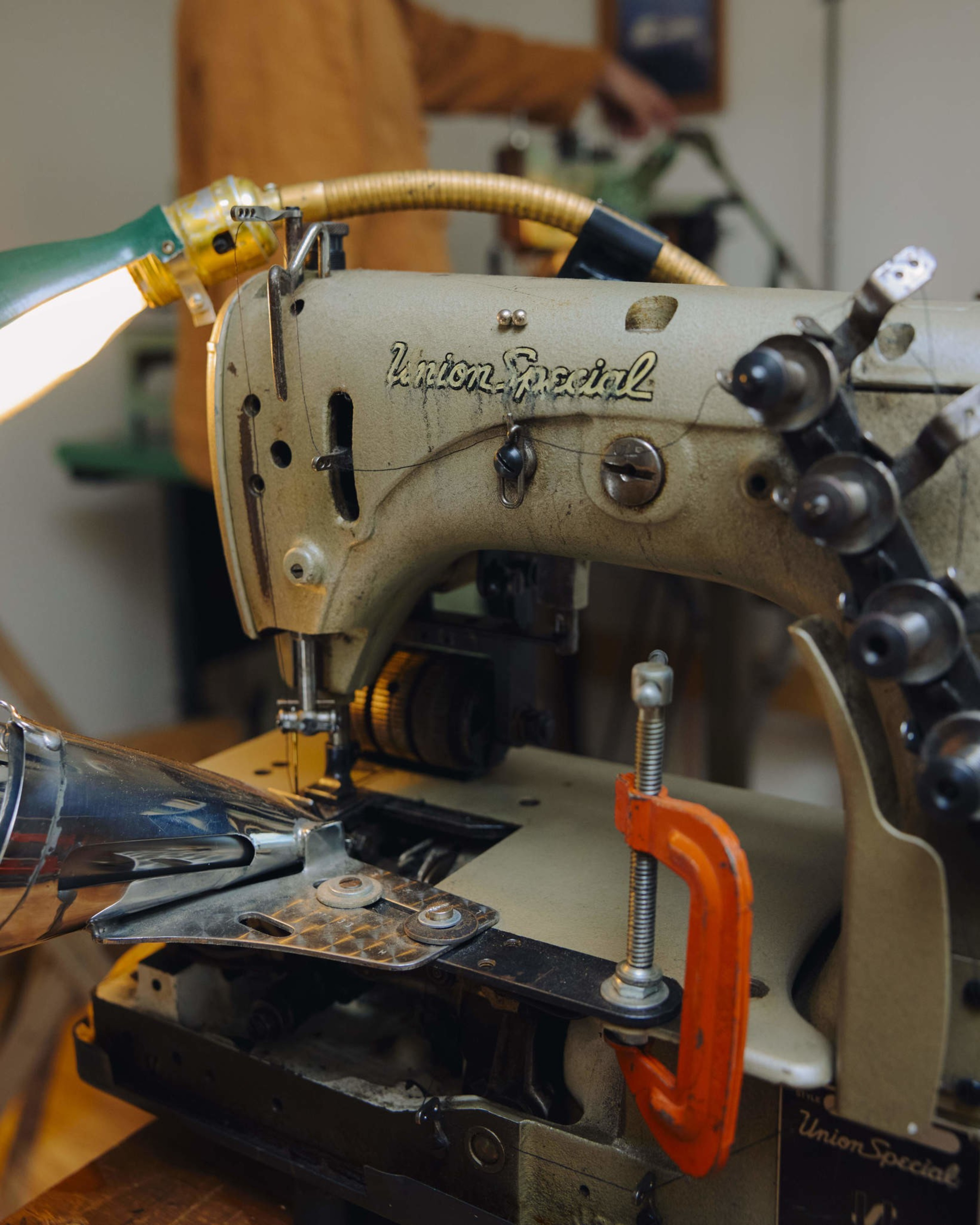 An old Union Special sewing machine with visible wear and thread, a bright orange clamp attached, and a flexible lamp illuminating the work area.