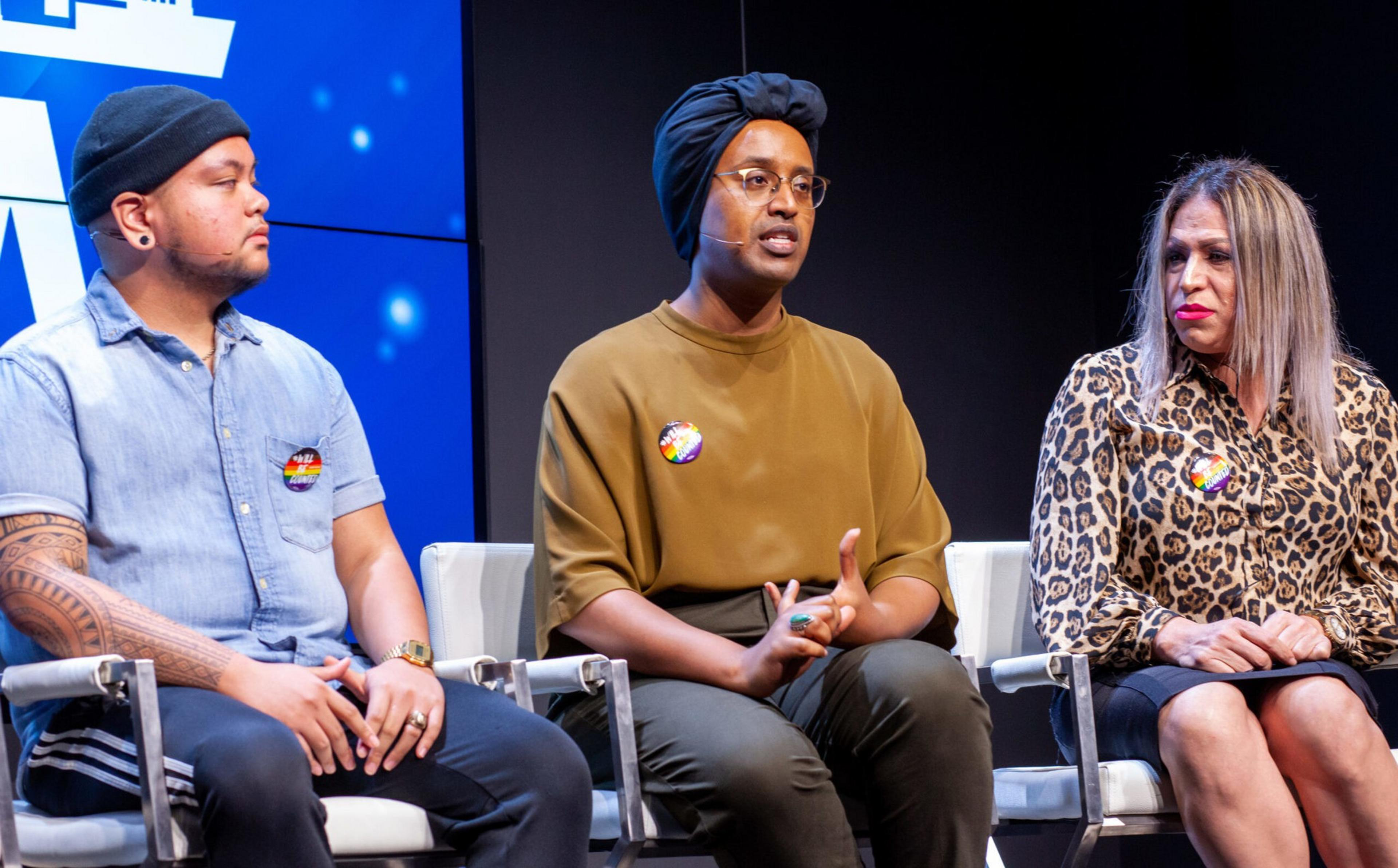Three people sit on stage chairs in conversation; one wears a turban and glasses, another sports tattoos and a beanie, and the third wears a leopard print blouse.