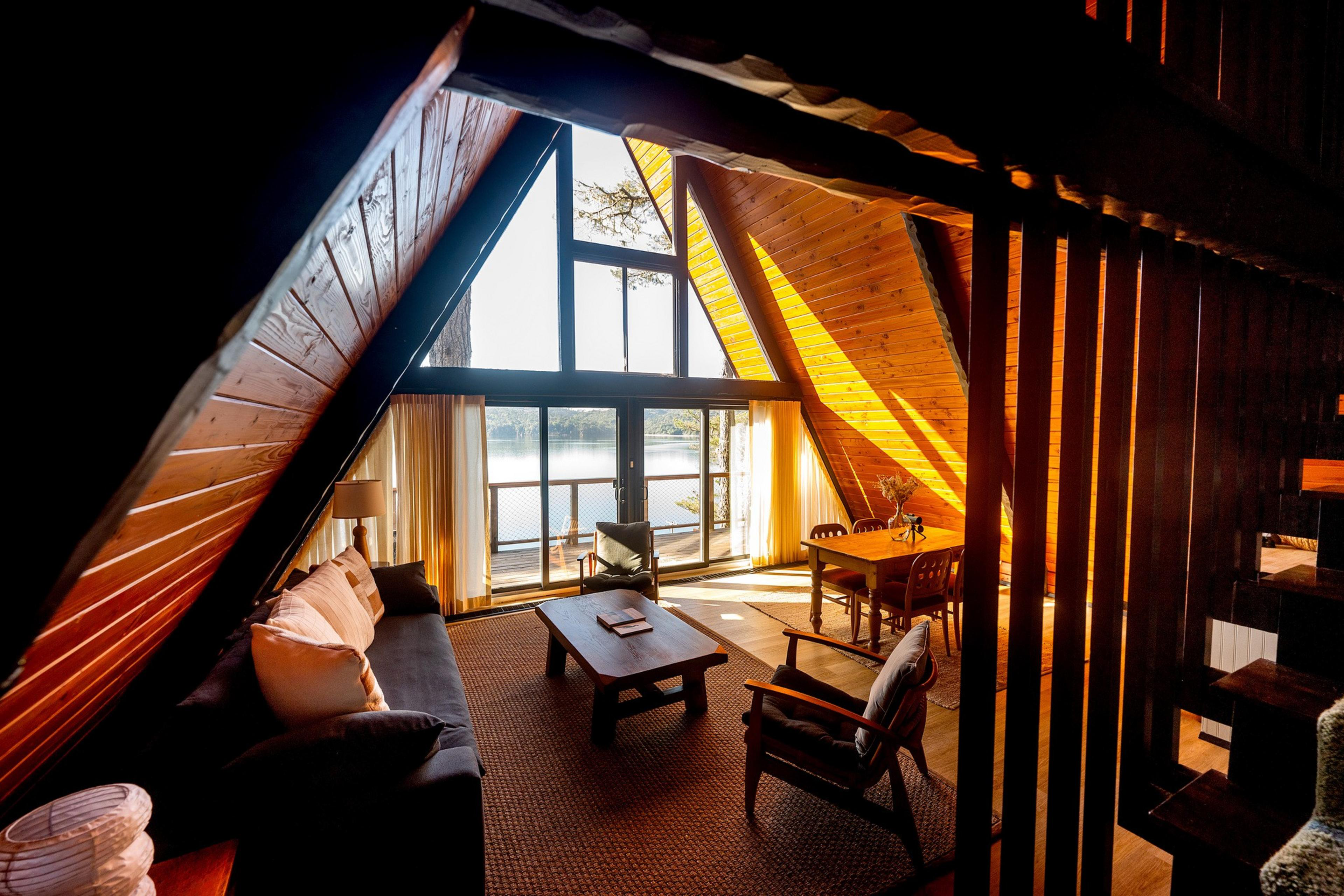 This image shows a cozy A-frame cabin interior with a sofa, armchairs, and a table. Large windows offer a stunning lake view, and sunlight streams in warmly.