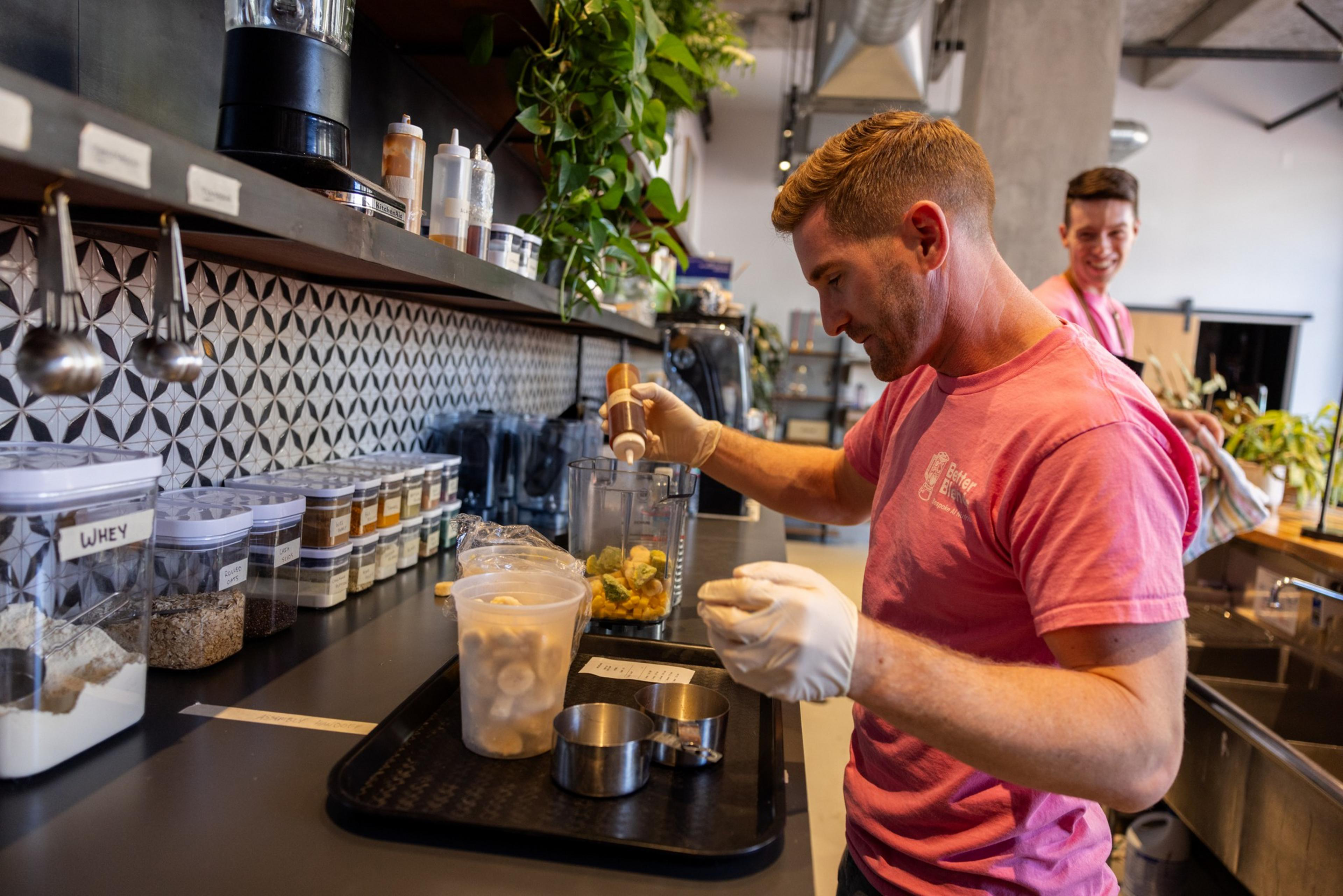 A man wearing a pink shirt and gloves adds an ingredient into a blender filled with fruit, while another person smiles in the background of a kitchen.