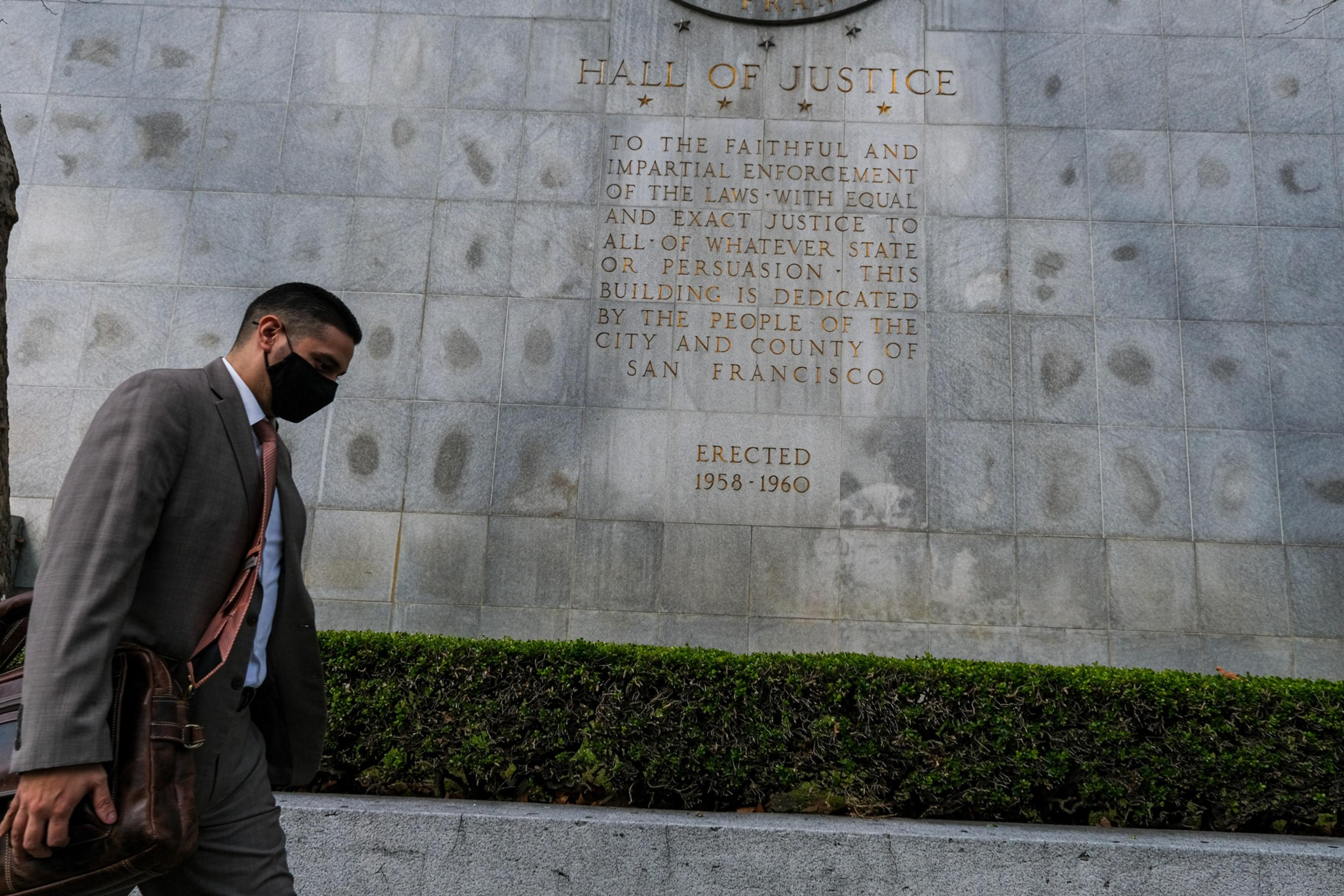 A man in a suit and mask walks past a wall engraved with the dedication of the Hall of Justice in San Francisco, erected 1958-1960.