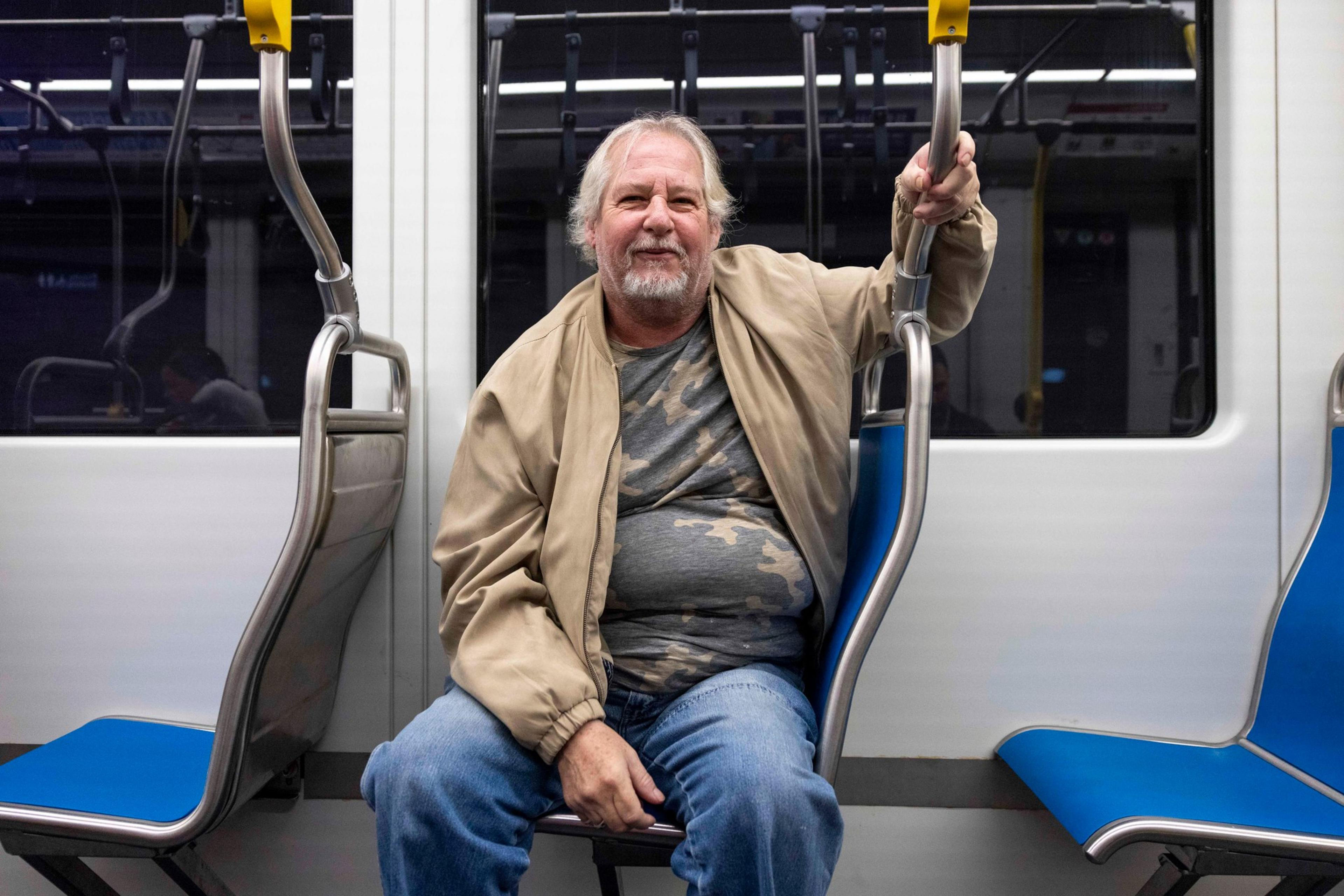 An older man with a beard, wearing a beige jacket and camouflage shirt, sits inside a train, holding the handrail above him.