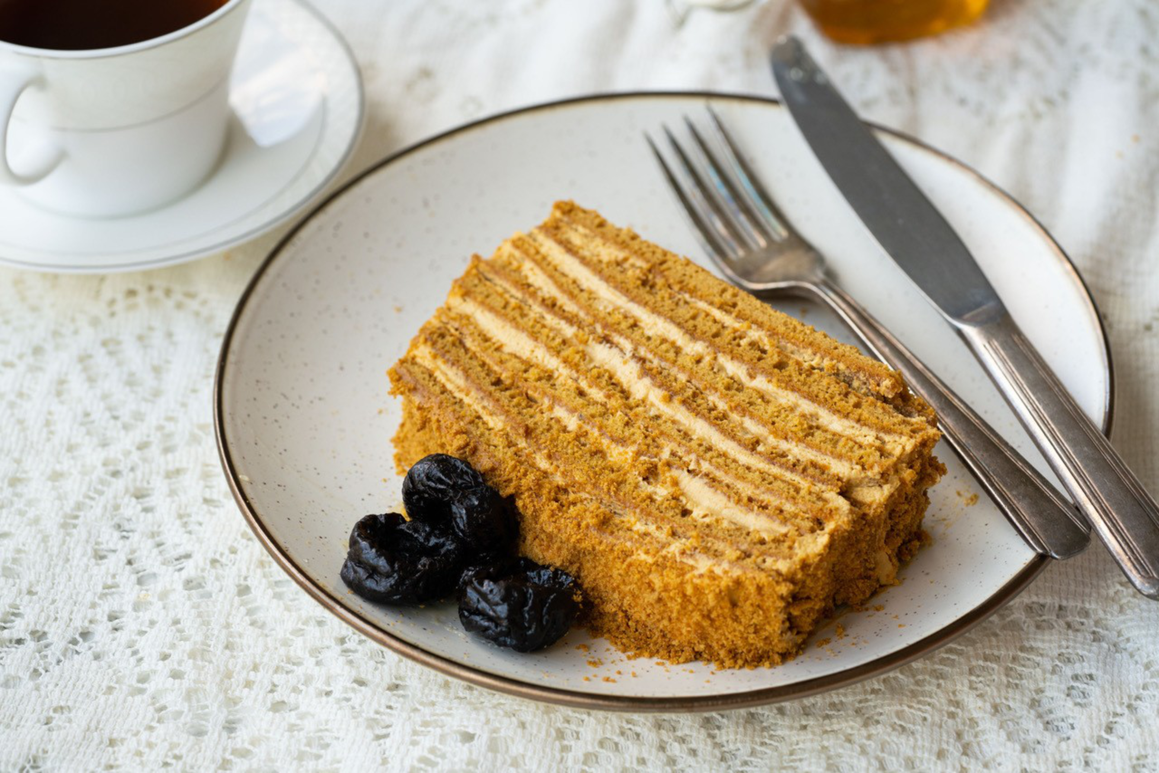 A layered honey cake slice with three prunes on a white plate, accompanied by a fork and knife, resting on a white lace tablecloth next to a cup of tea.