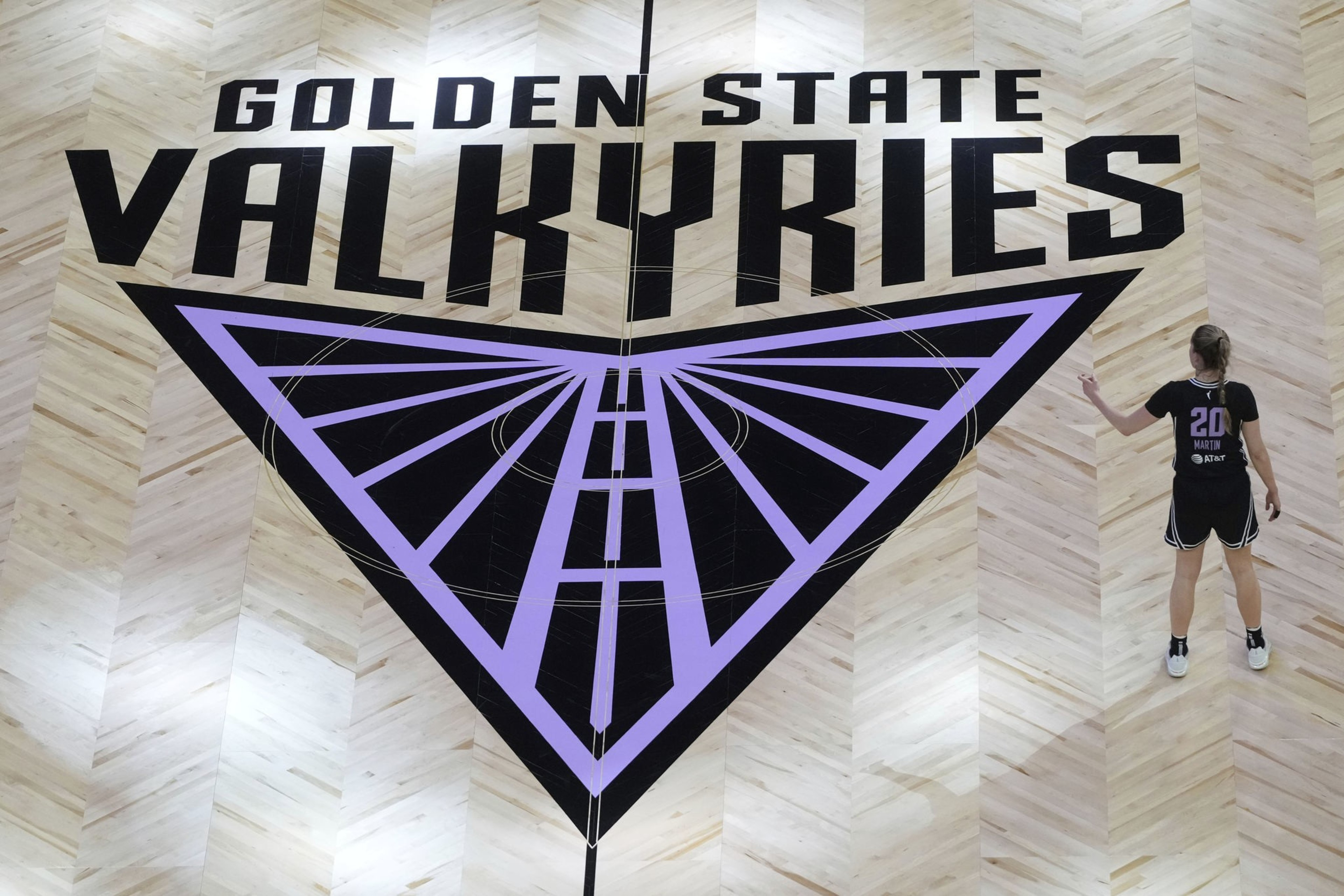 A wooden basketball court floor with a large purple and black Golden State Valkyries logo, and a player wearing a black jersey standing nearby.