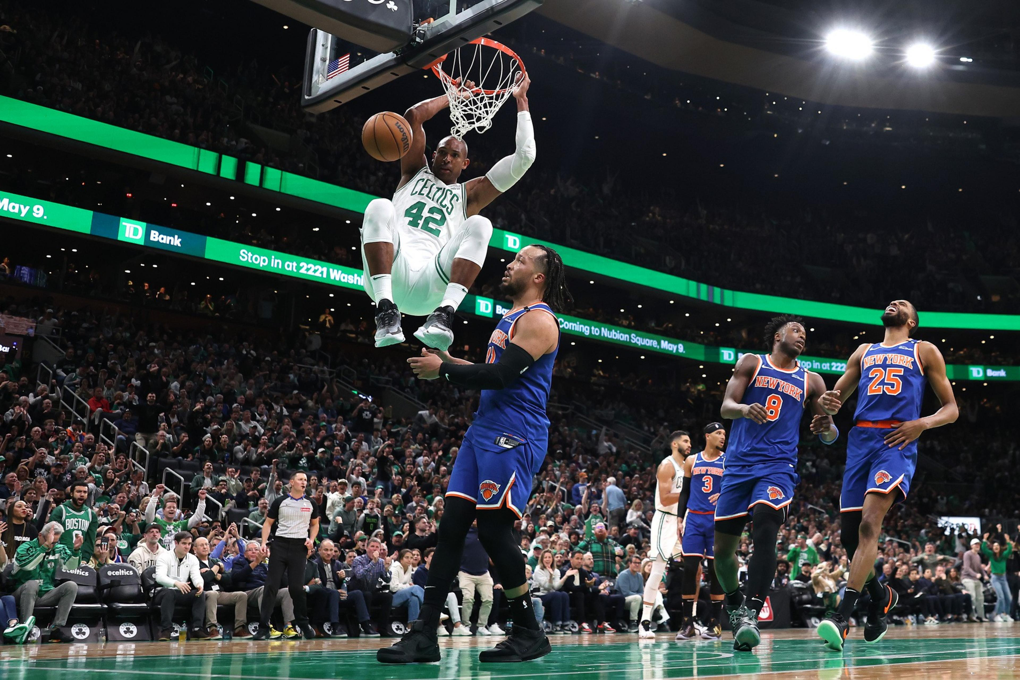 A Celtics player dunks with the ball near the basket as Knicks players watch, surrounded by a packed crowd in an NBA game.