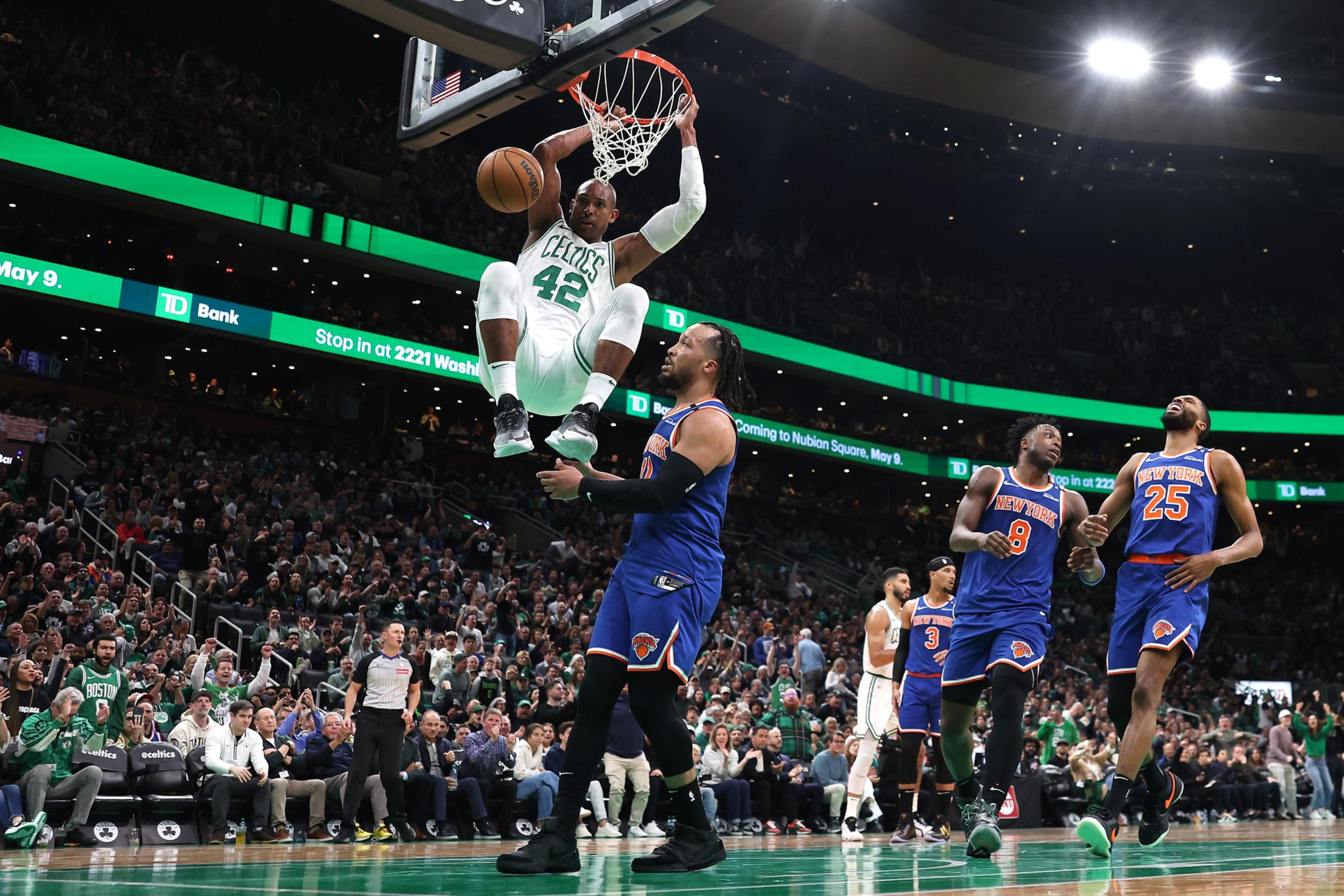 A Celtics player dunks with the ball near the basket as Knicks players watch, surrounded by a packed crowd in an NBA game.