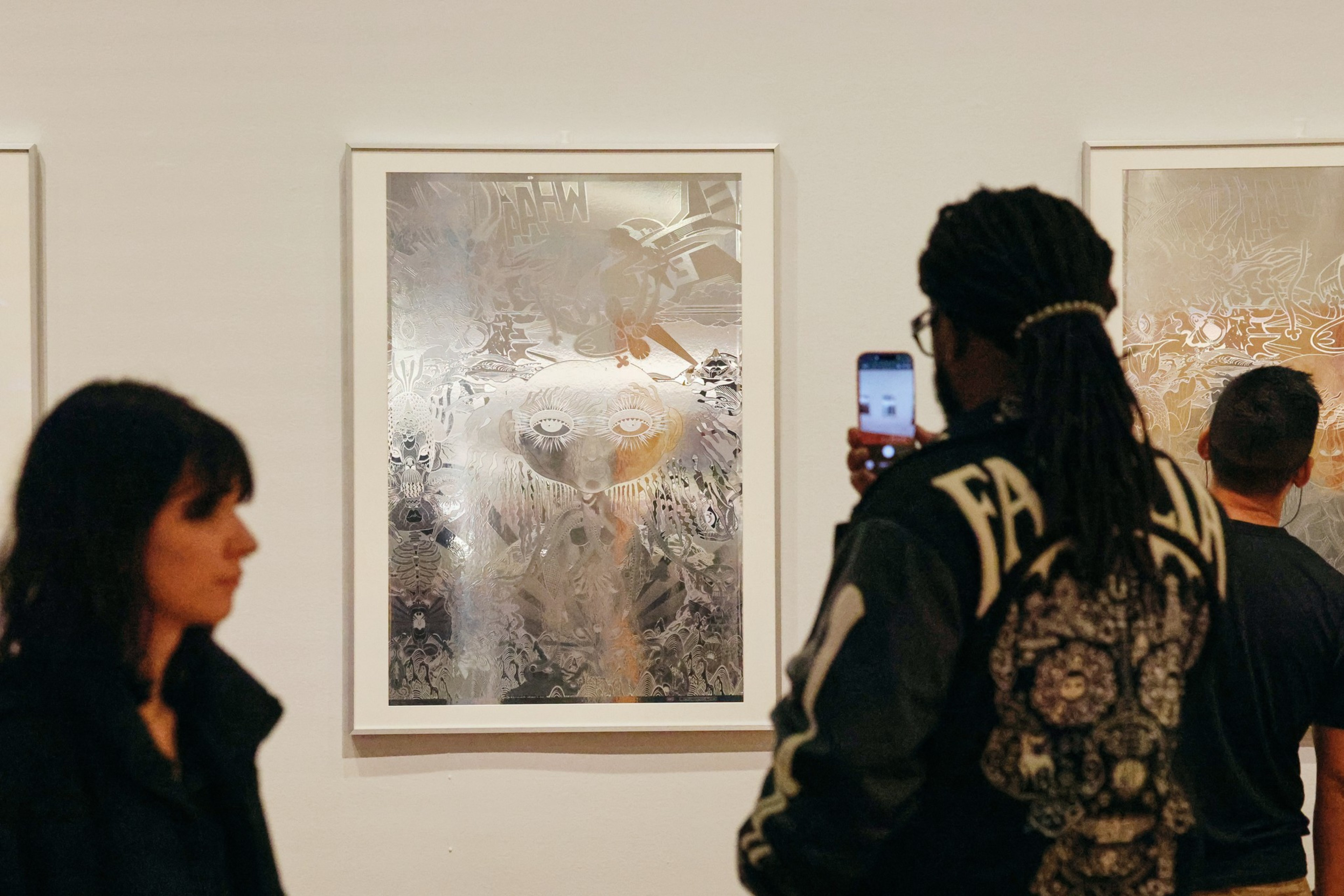 Two framed abstract artworks hang on a white wall as three people observe; one person uses a phone to photograph an artwork in the center.