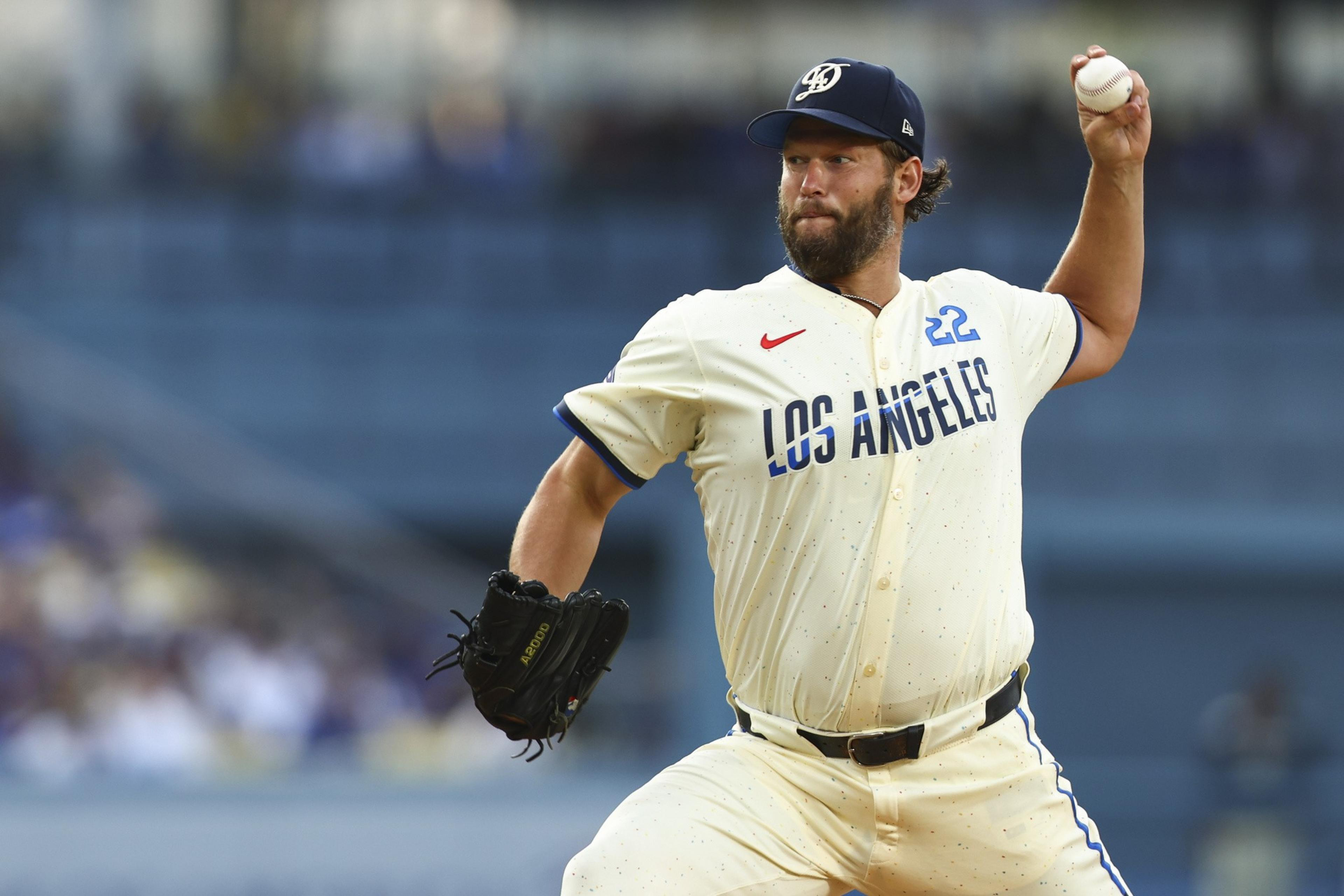 A baseball player is pitching in a game. He is wearing a cream-colored uniform with &quot;Los Angeles&quot; on the front and a navy cap, holding a baseball in one hand.