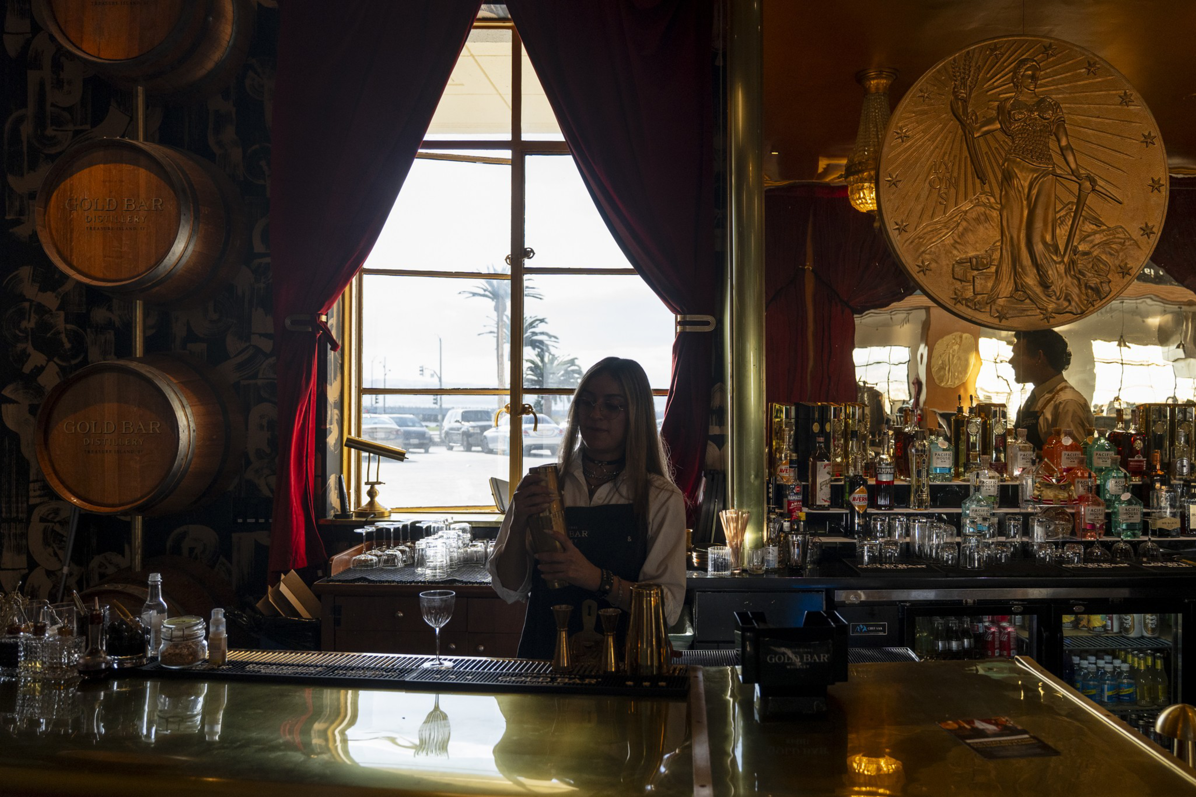 A bartender shakes a cocktail behind a bar with wooden barrels, glassware, and liquor bottles, lit by natural light from a window with red curtains.