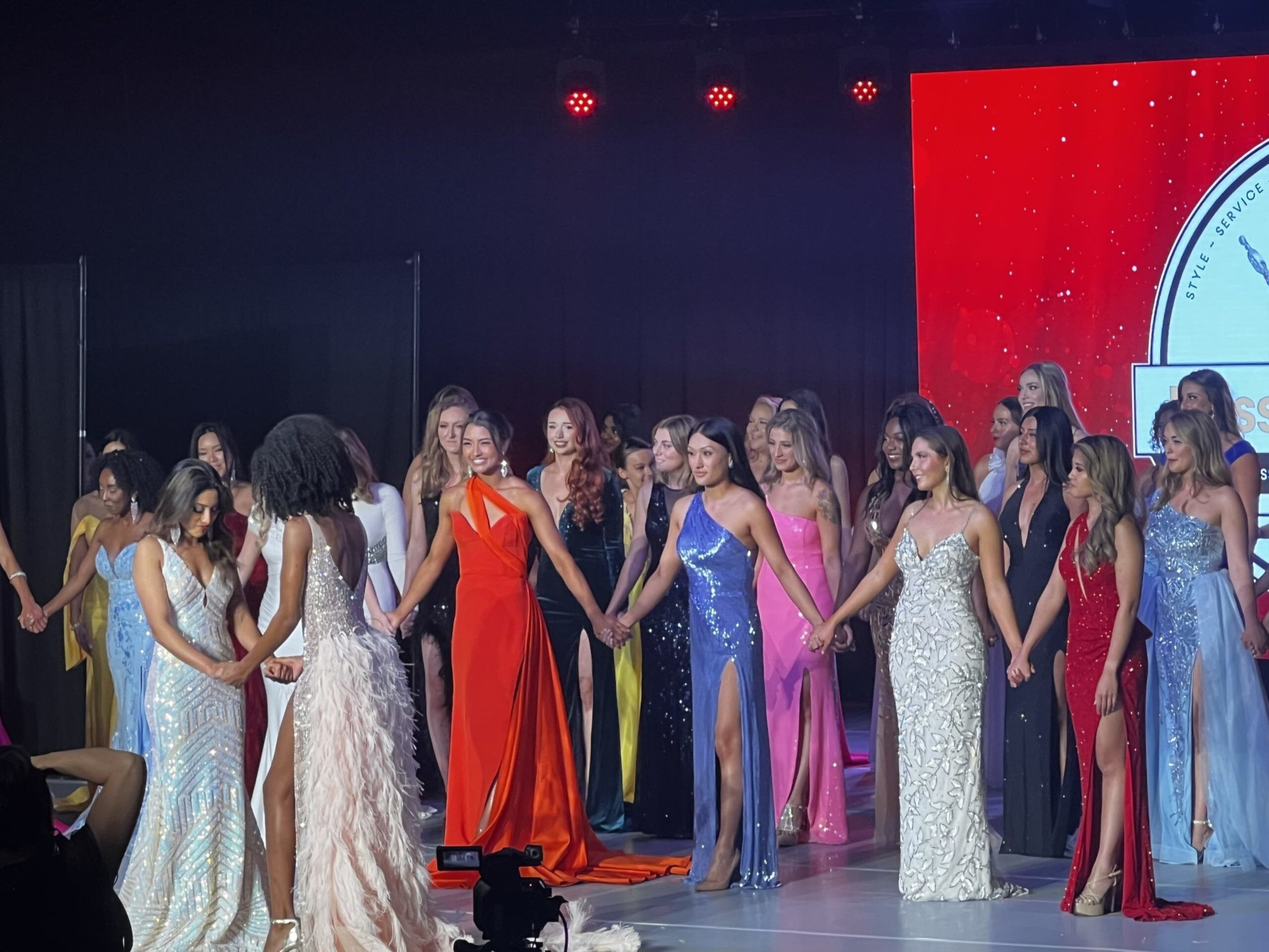 Women in various elegant evening gowns hold hands on a stage, illuminated by red lights, with a large red screen visible in the background.