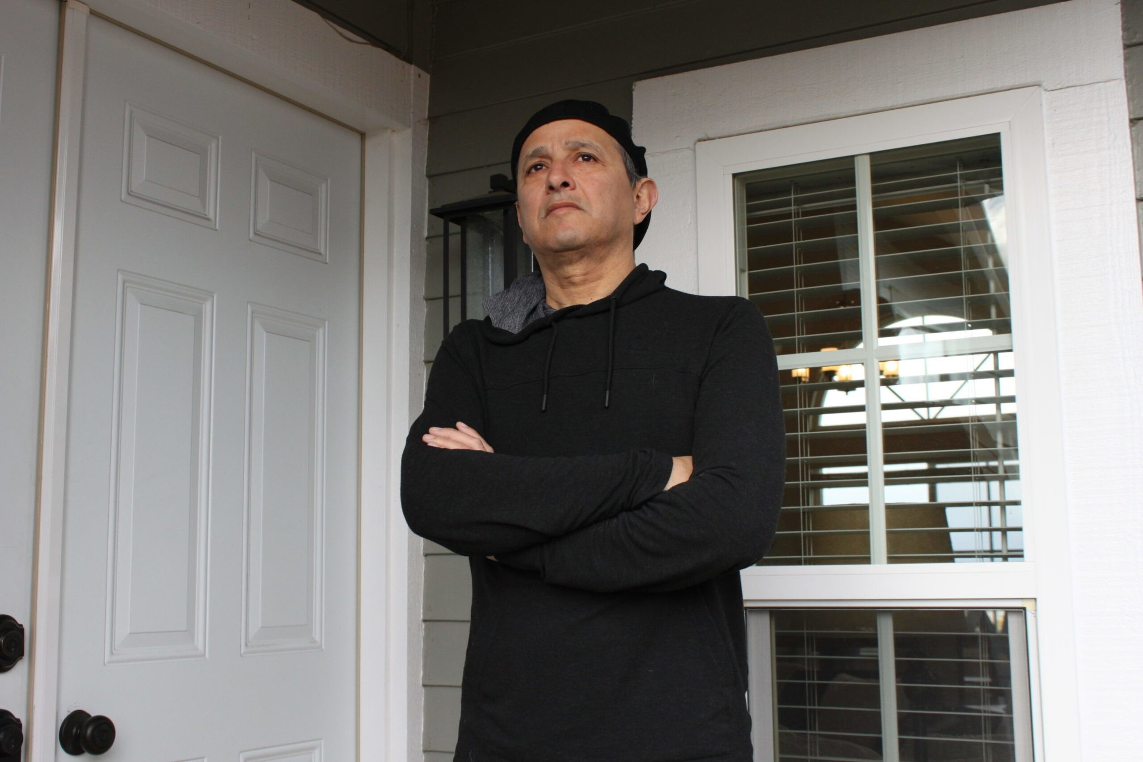 A man in a black hoodie and hat stands with arms crossed outside by a white door and a window with blinds, looking off to the side.