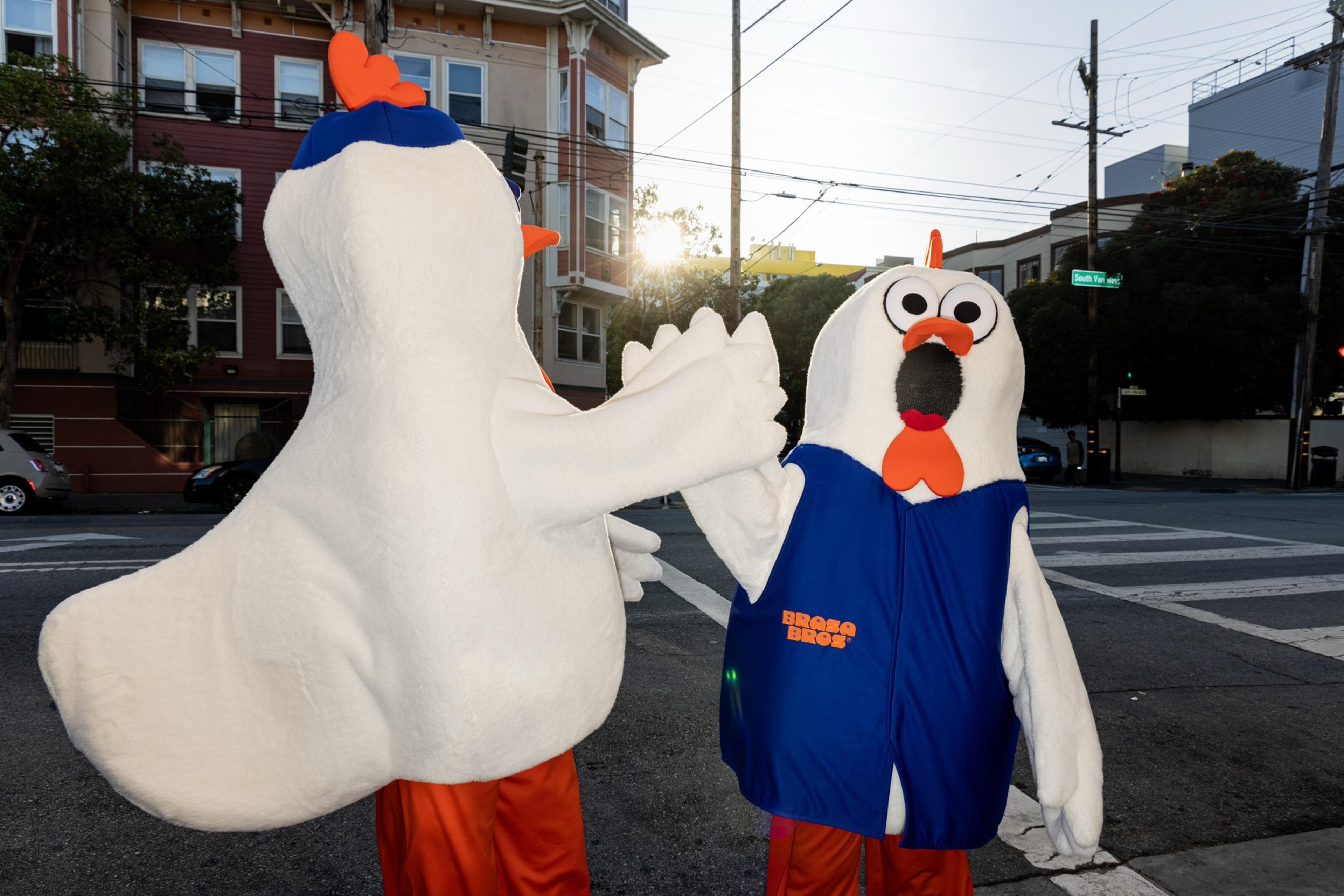 Two people in large white chicken costumes with orange beaks and feet give each other a high-five on a city street corner at sunset.