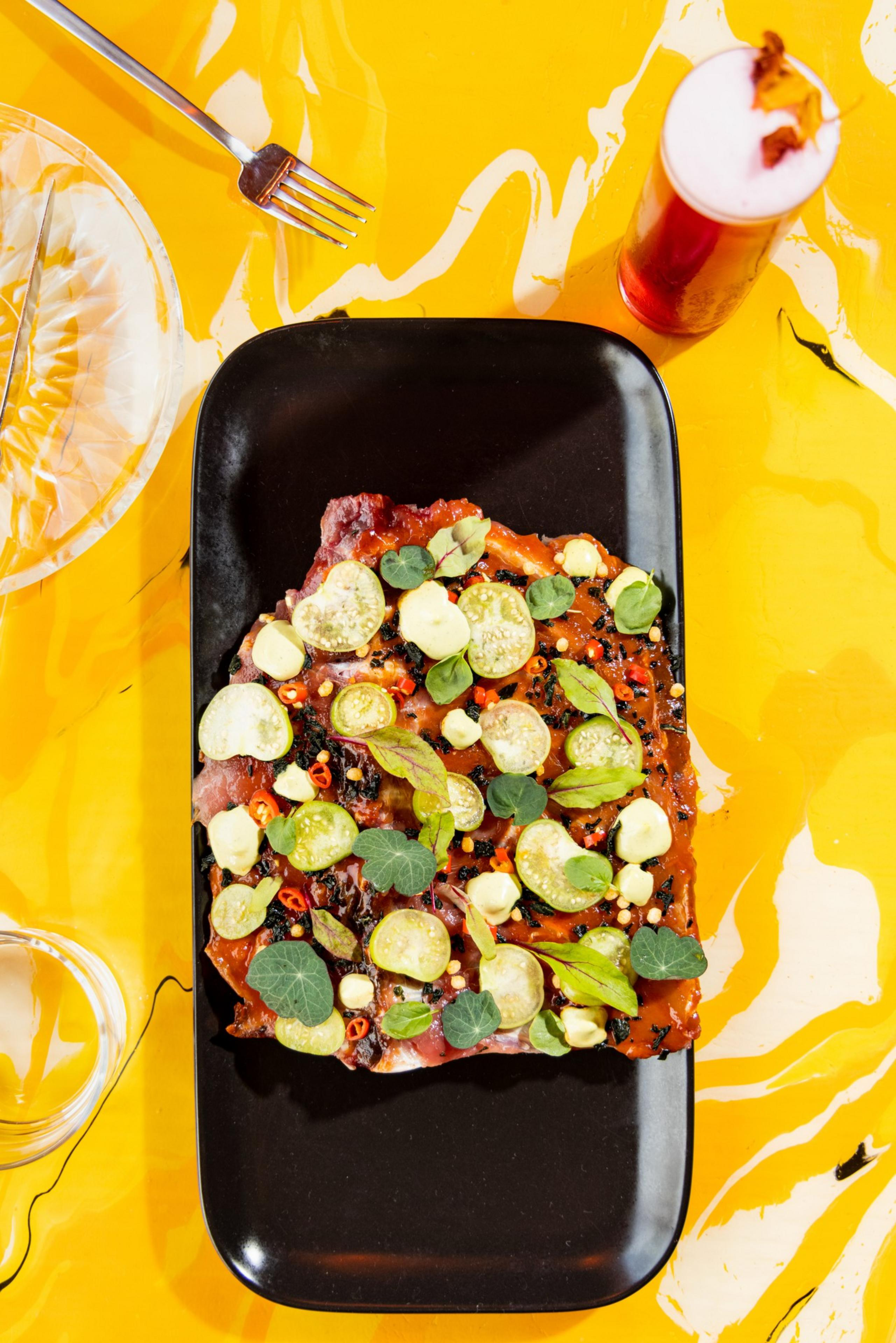 A rectangular black plate holds a piece of food topped with sliced green pickles, small dollops of white sauce, red chili slices, and green leaves on a yellow marbled surface.