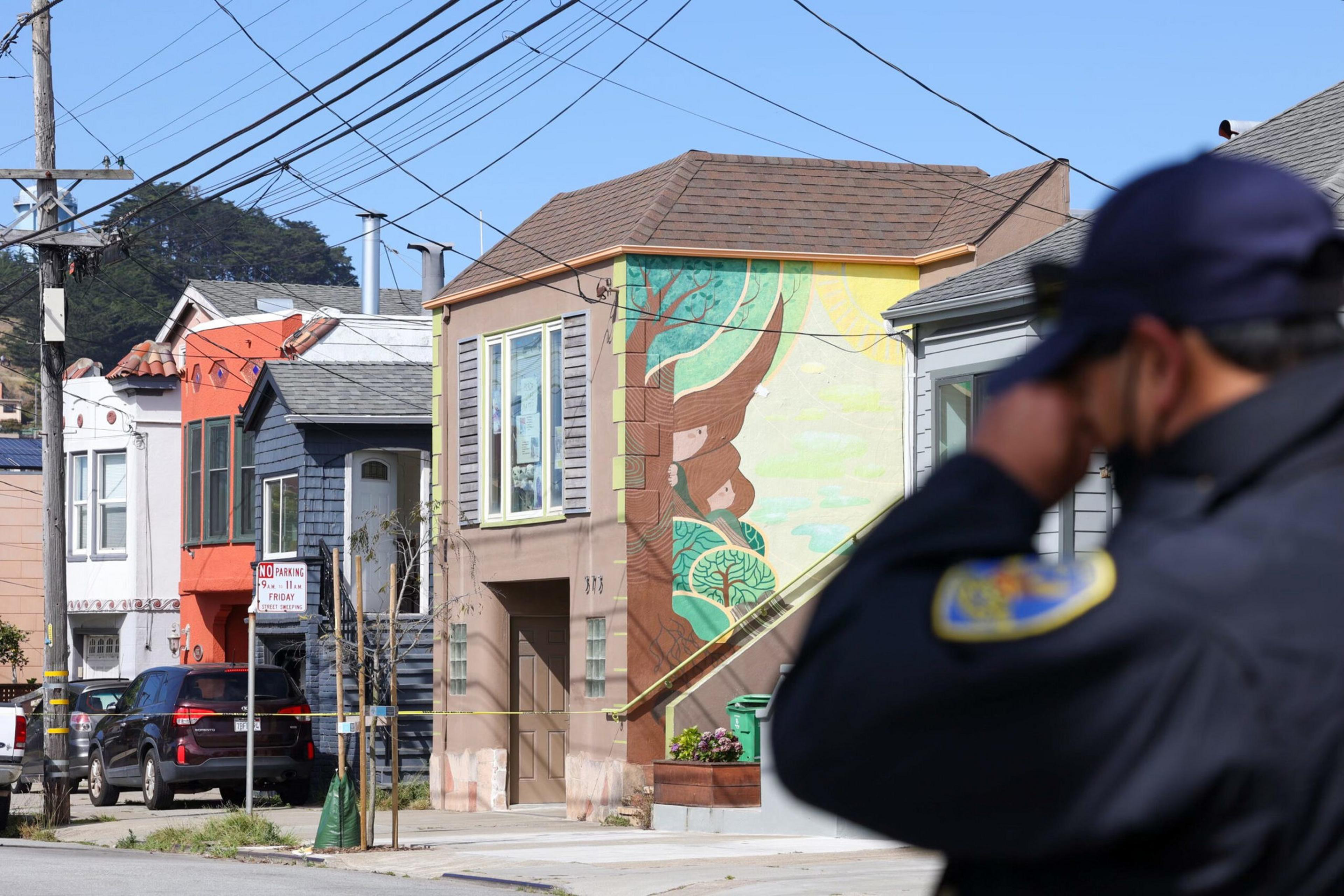 A police officer in uniform looks toward three houses, one with a colorful tree mural, with several parked cars and a clear blue sky overhead.