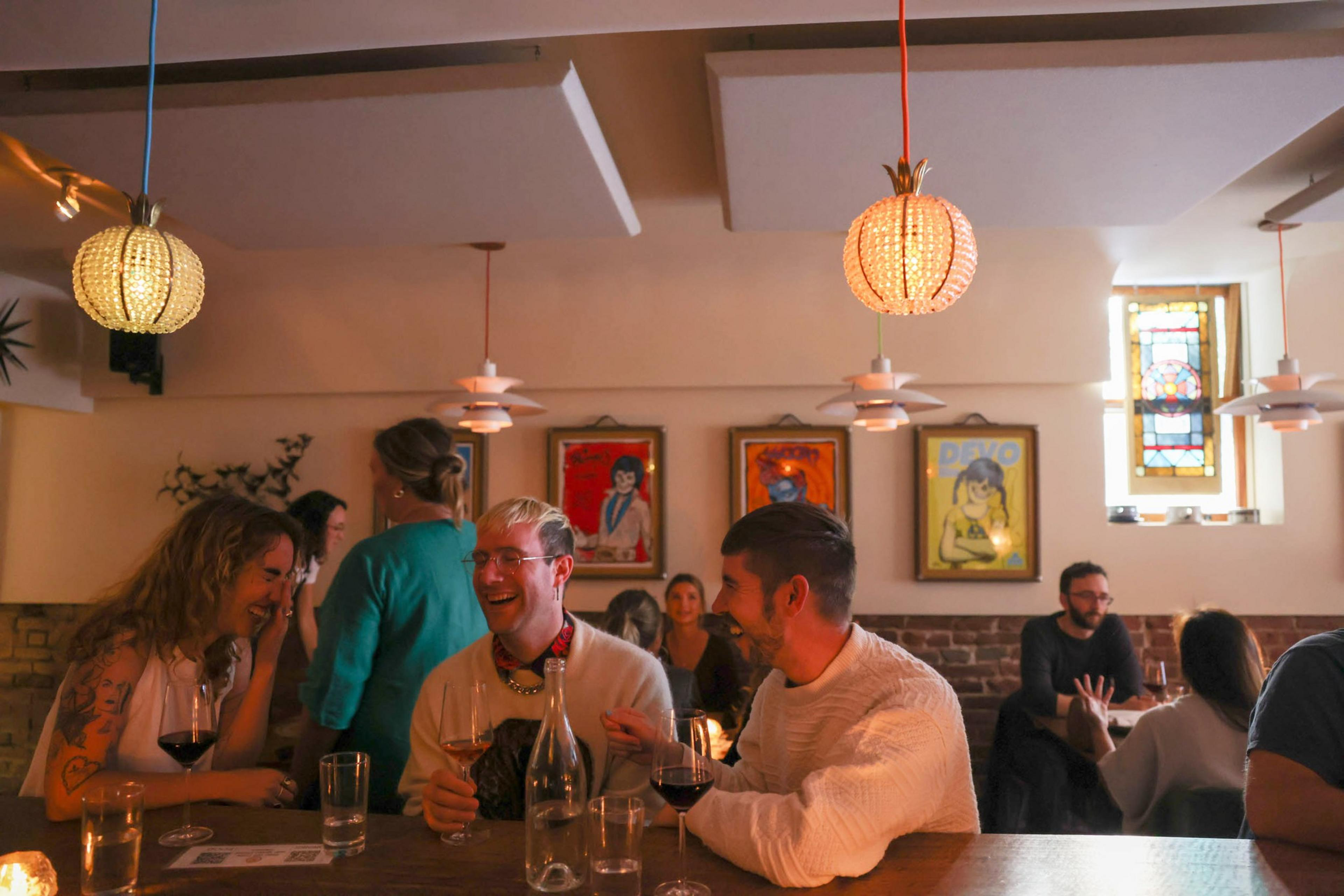 Three people laugh and chat over wine at a cozy bar with warm lighting, framed artwork, and a stained glass window in the background.