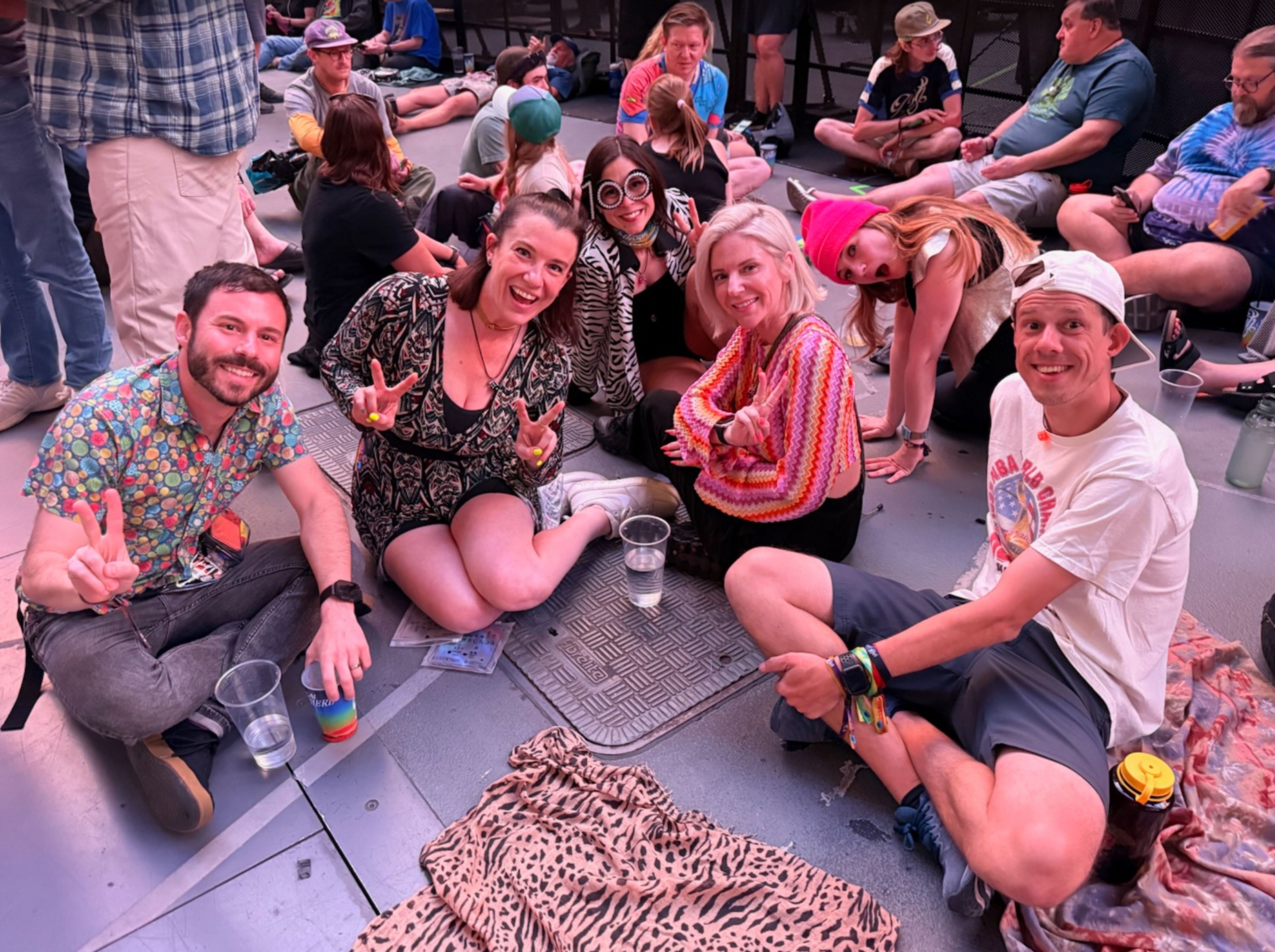 A group of six people sit on the floor smiling and making peace signs, surrounded by others also seated in a casual, lively setting.