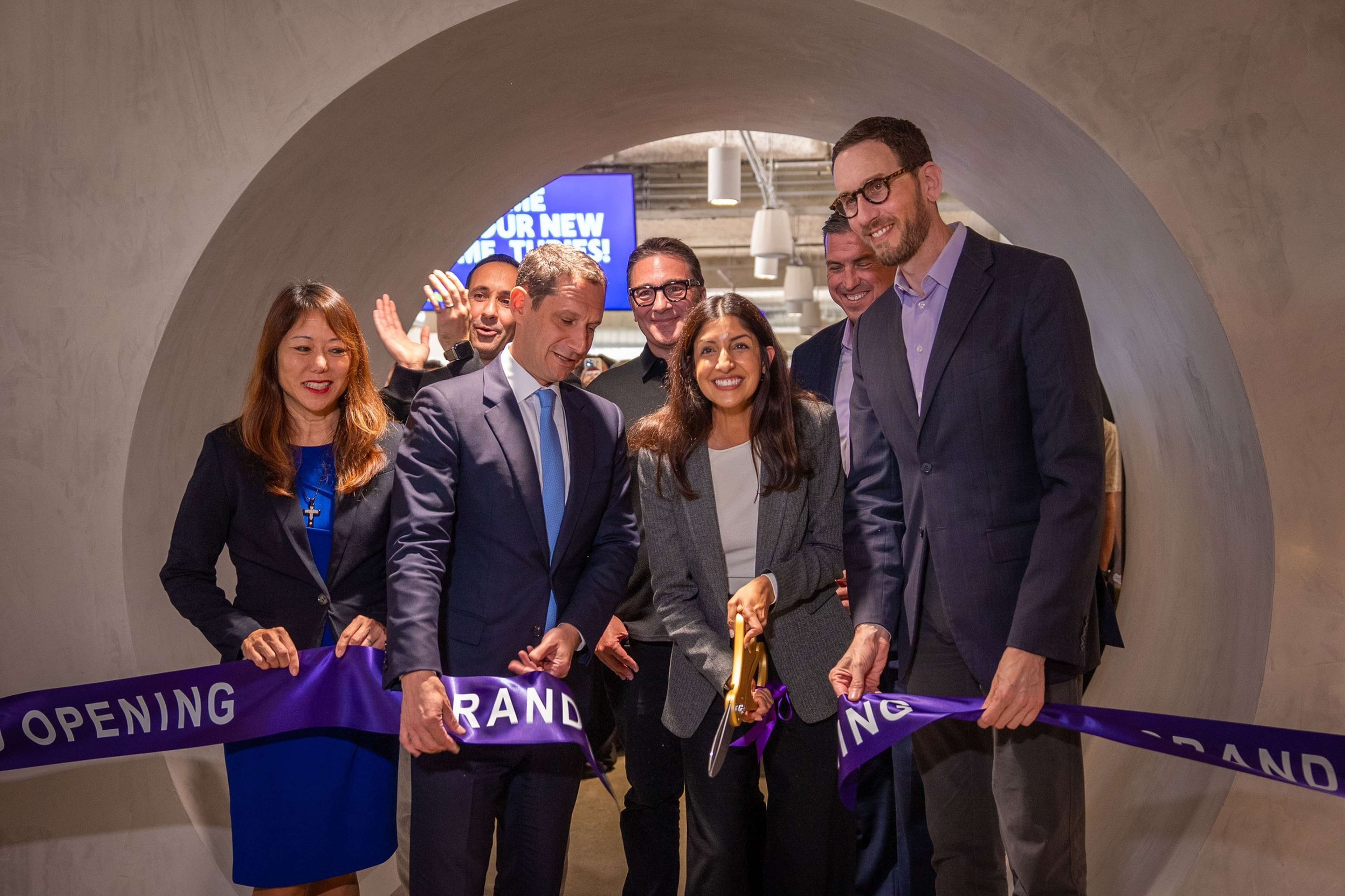 A group of seven well-dressed people smile as a woman cuts a purple ribbon at a grand opening event inside a modern, curved tunnel entrance.