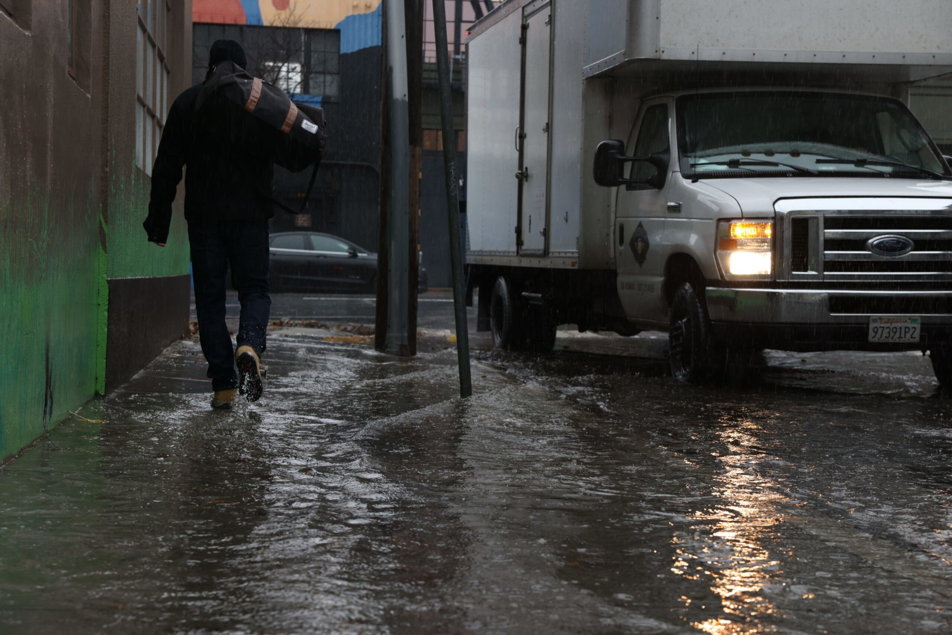 A person walks on a flooded street wearing dark clothes, carrying a bag, while a white truck with headlights on waits nearby in the rain.