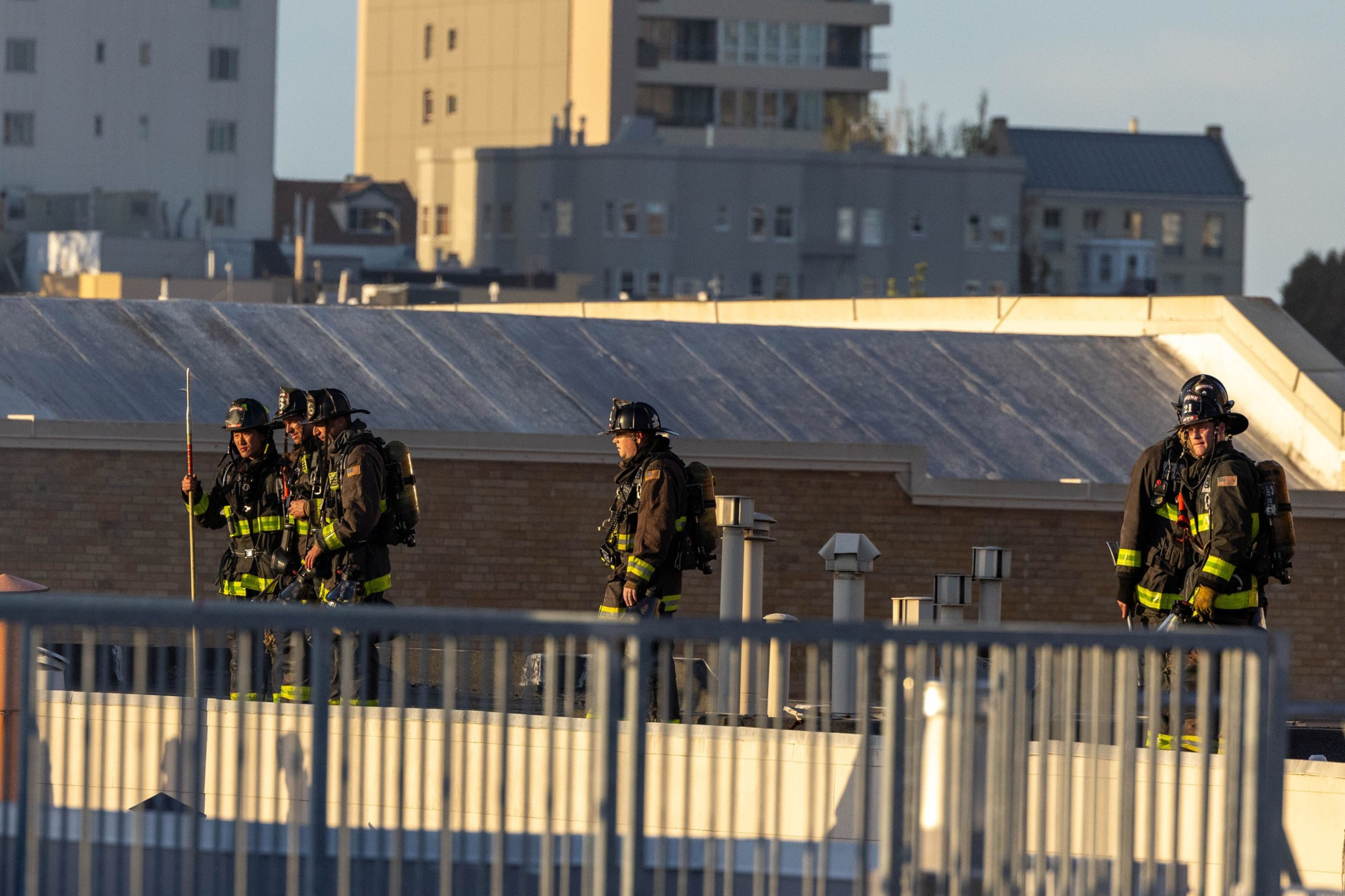 Four firefighters in full gear stand on a rooftop with urban buildings in the background, illuminated by sunlight.