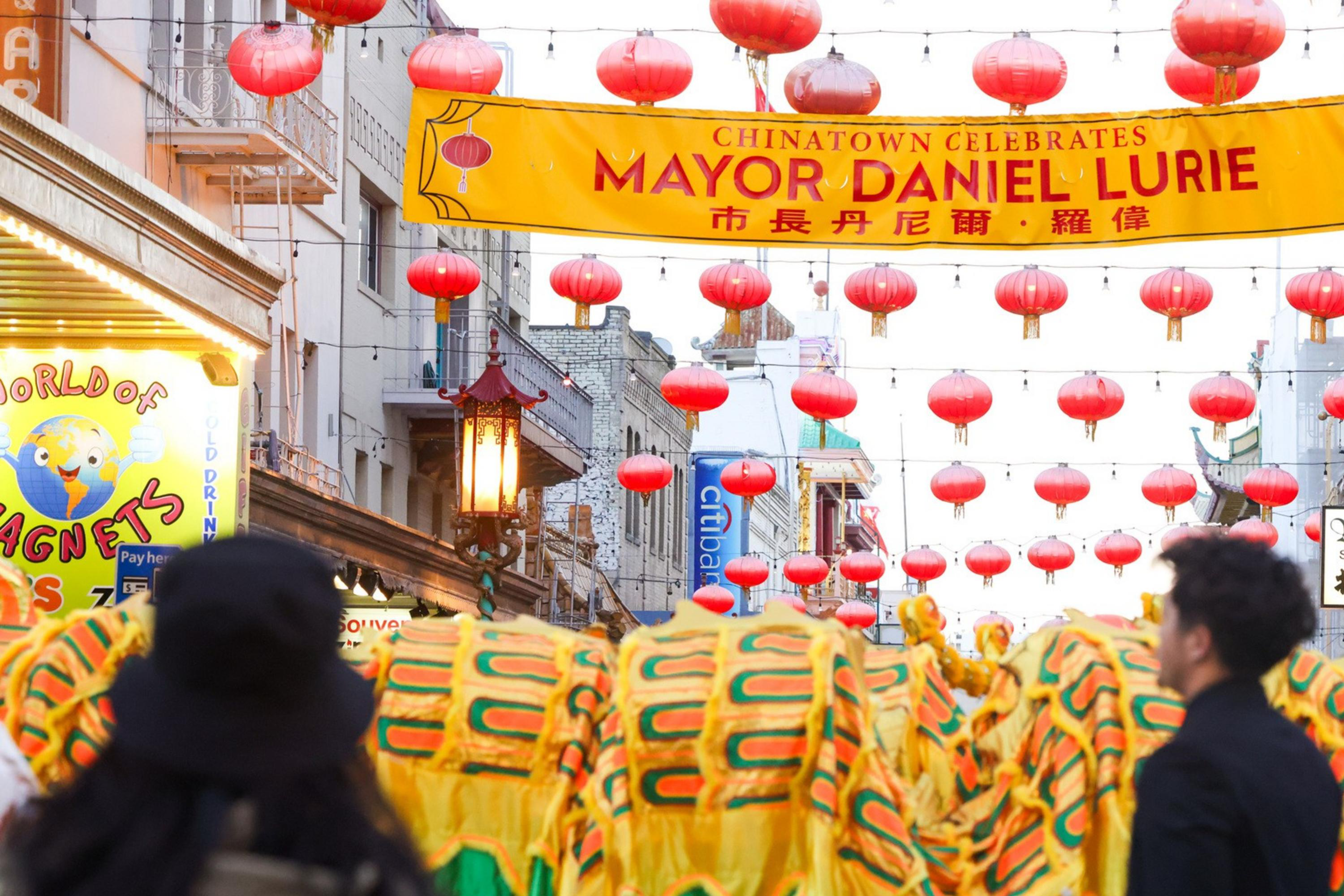 A vibrant street scene features a colorful dragon dance under red lanterns and a yellow banner celebrating Mayor Daniel Lurie in Chinatown.