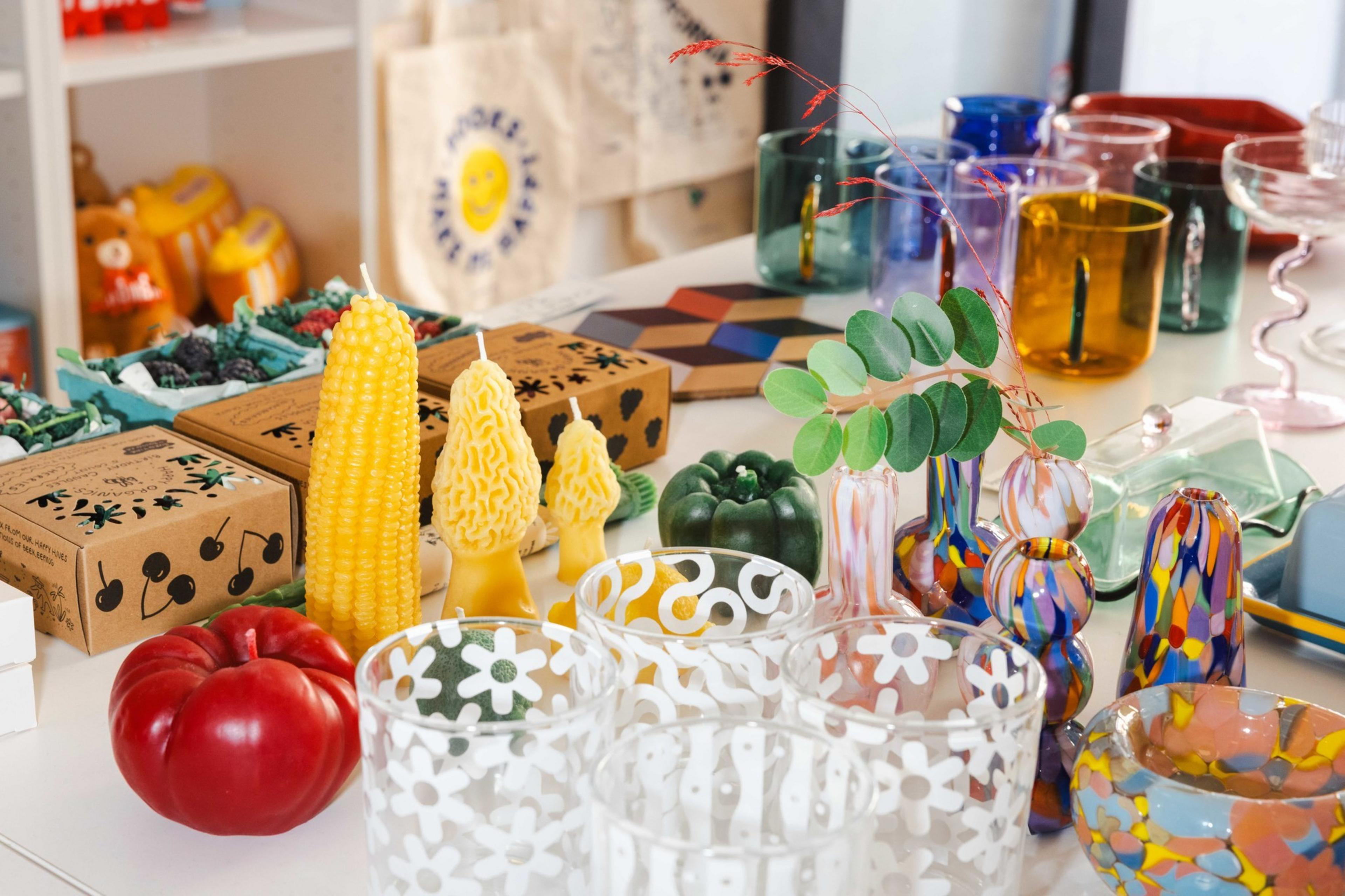 The image shows a variety of colorful glassware, decorative candles shaped like corn and trees, a red apple, and boxes with designs on a table.