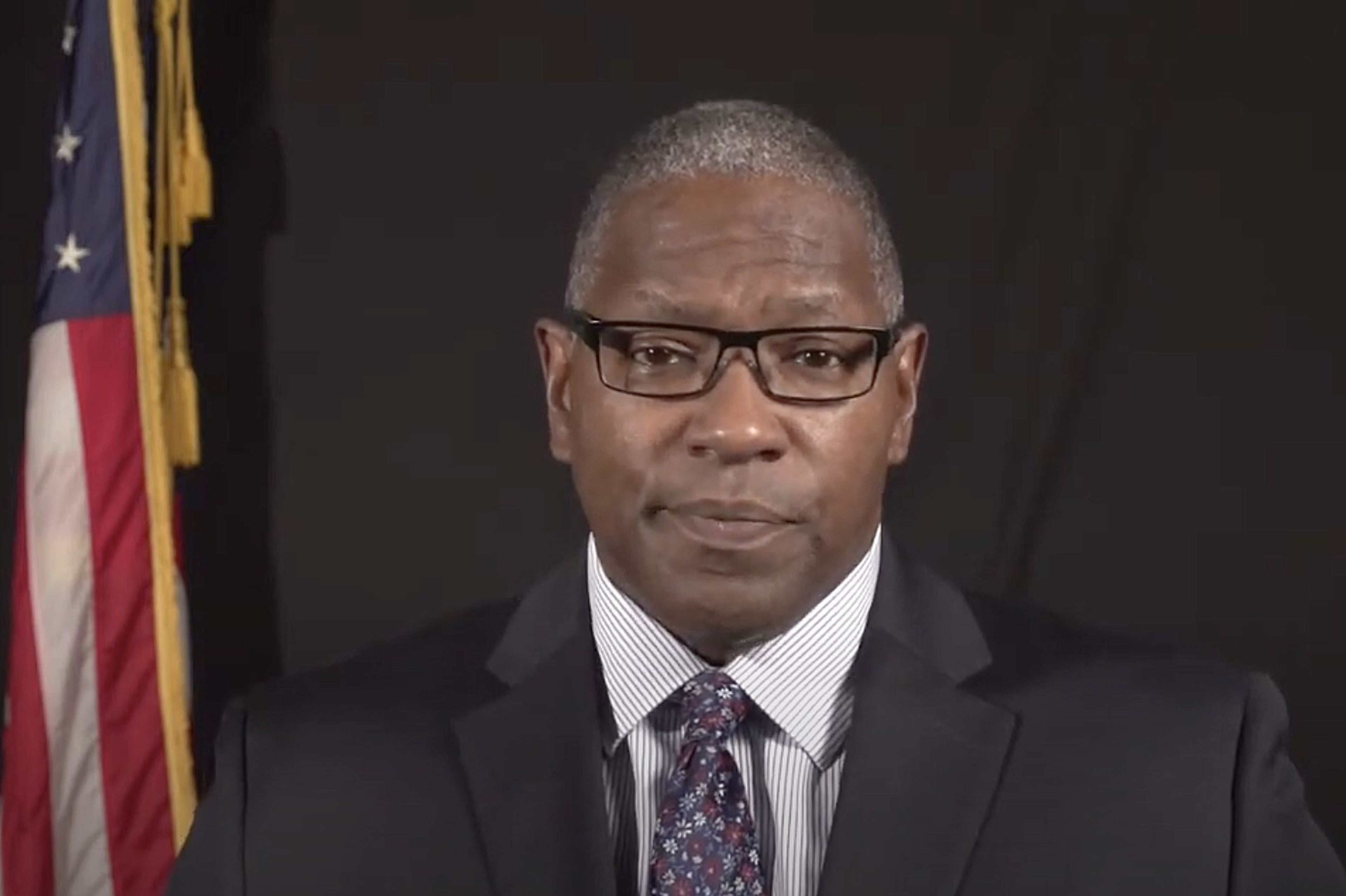 A man in a suit looks at the camera with the American flag in the background