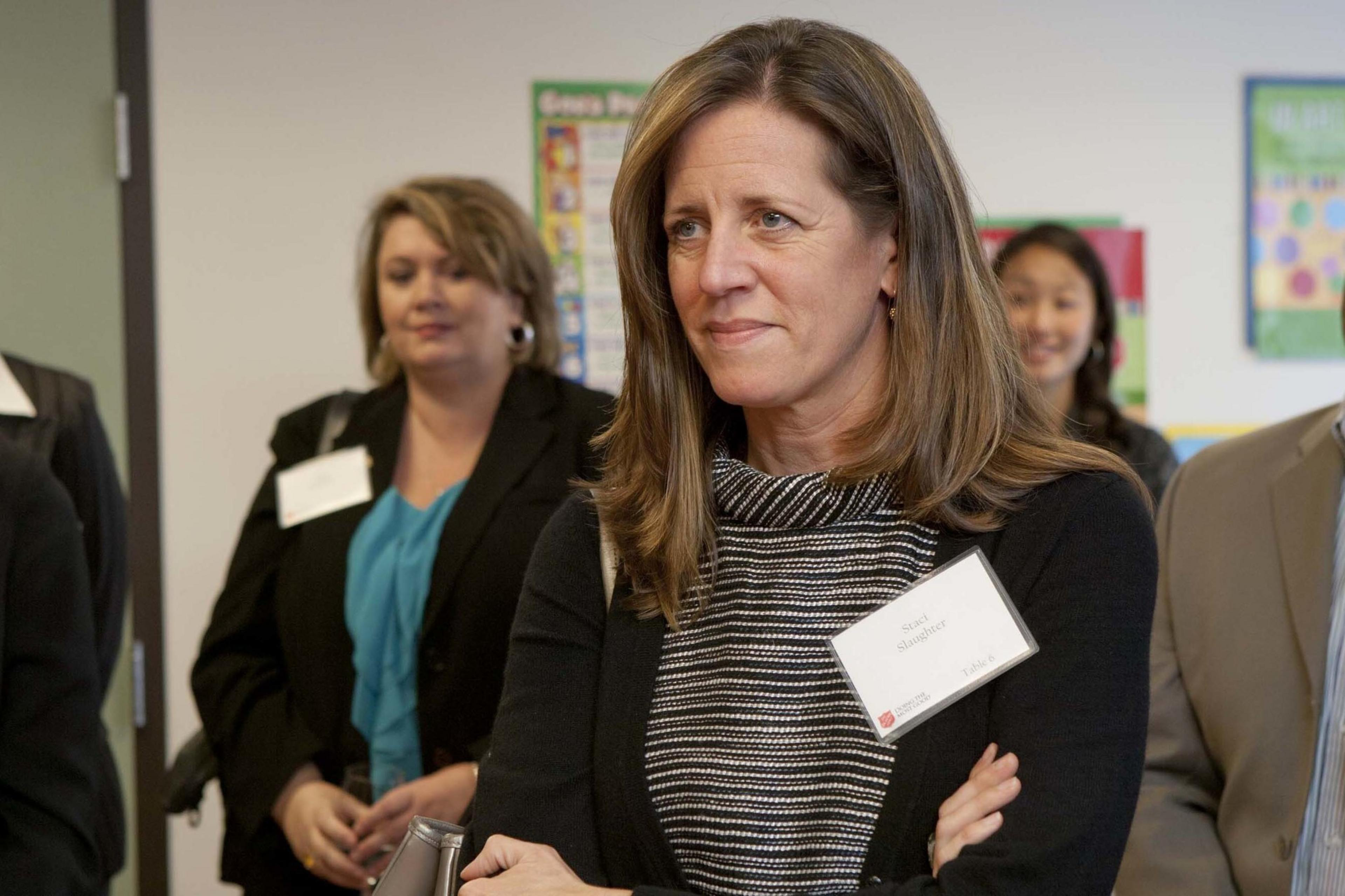 A woman with brown hair wearing a striped top stands with arms crossed, holding a nametag. She is in a group of adults, some of whom are smiling.