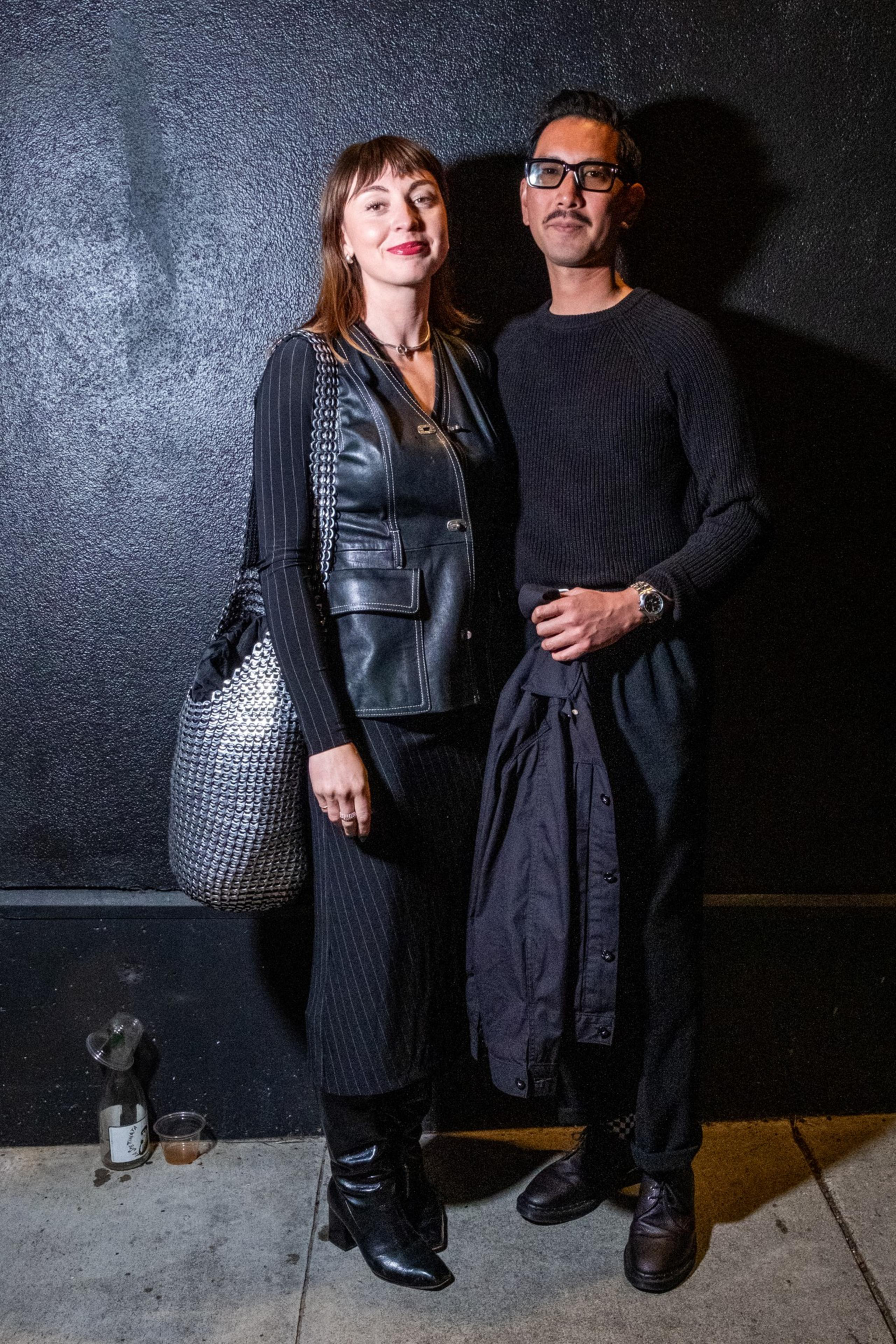 A woman and man stand against a dark wall. She wears a leather vest and carries a shiny bag, while he is in a black sweater holding a jacket.