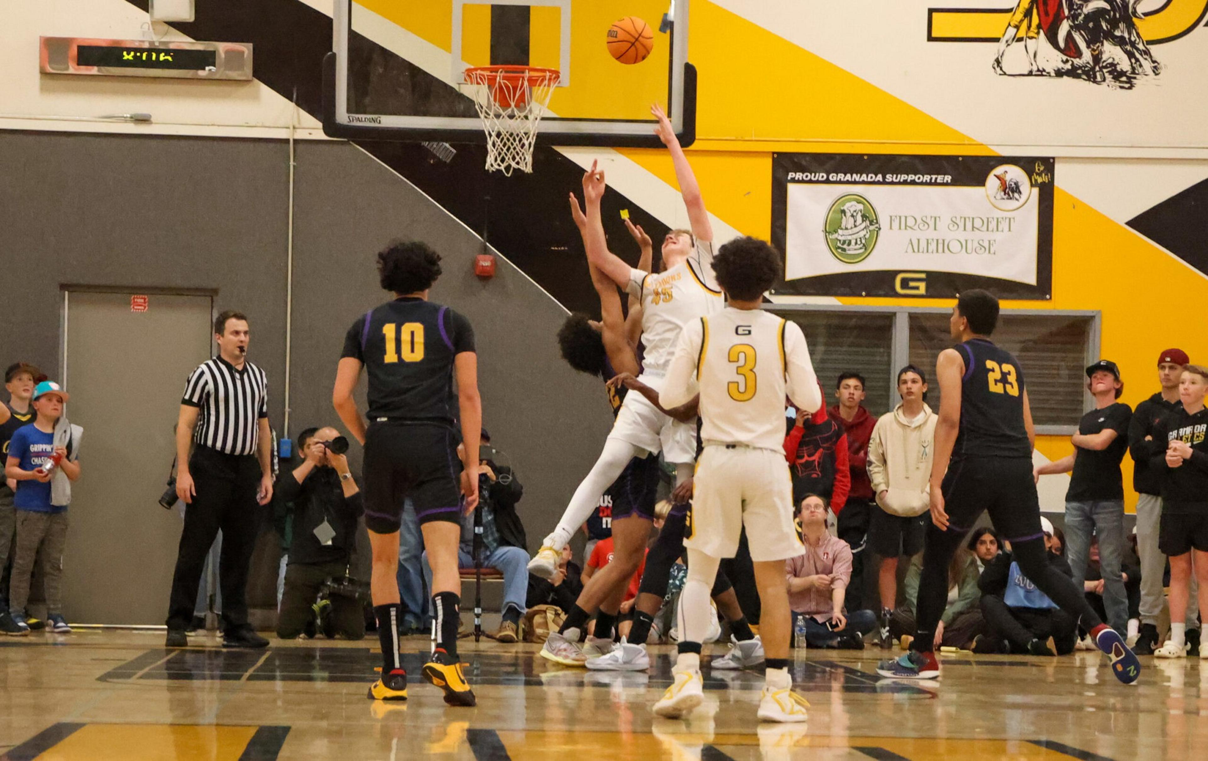 A basketball player in white jumps to shoot while defenders in black and yellow attempt to block, with spectators and a referee watching nearby.