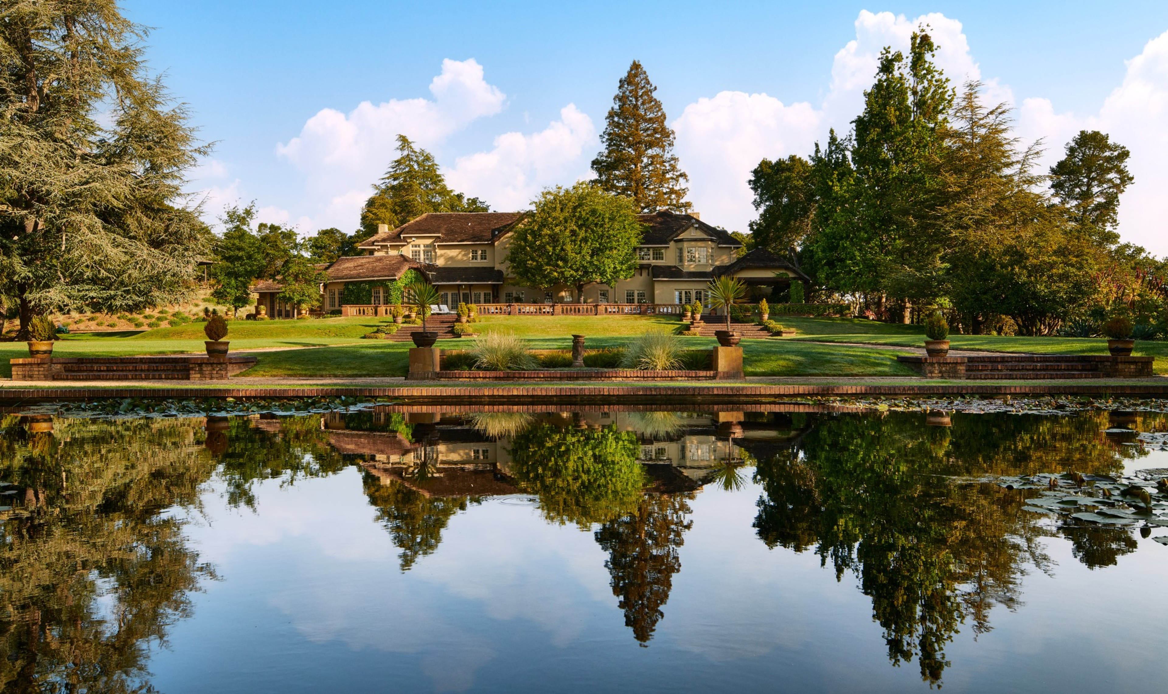 A mansion sits on a grassy hill with a pond in front.