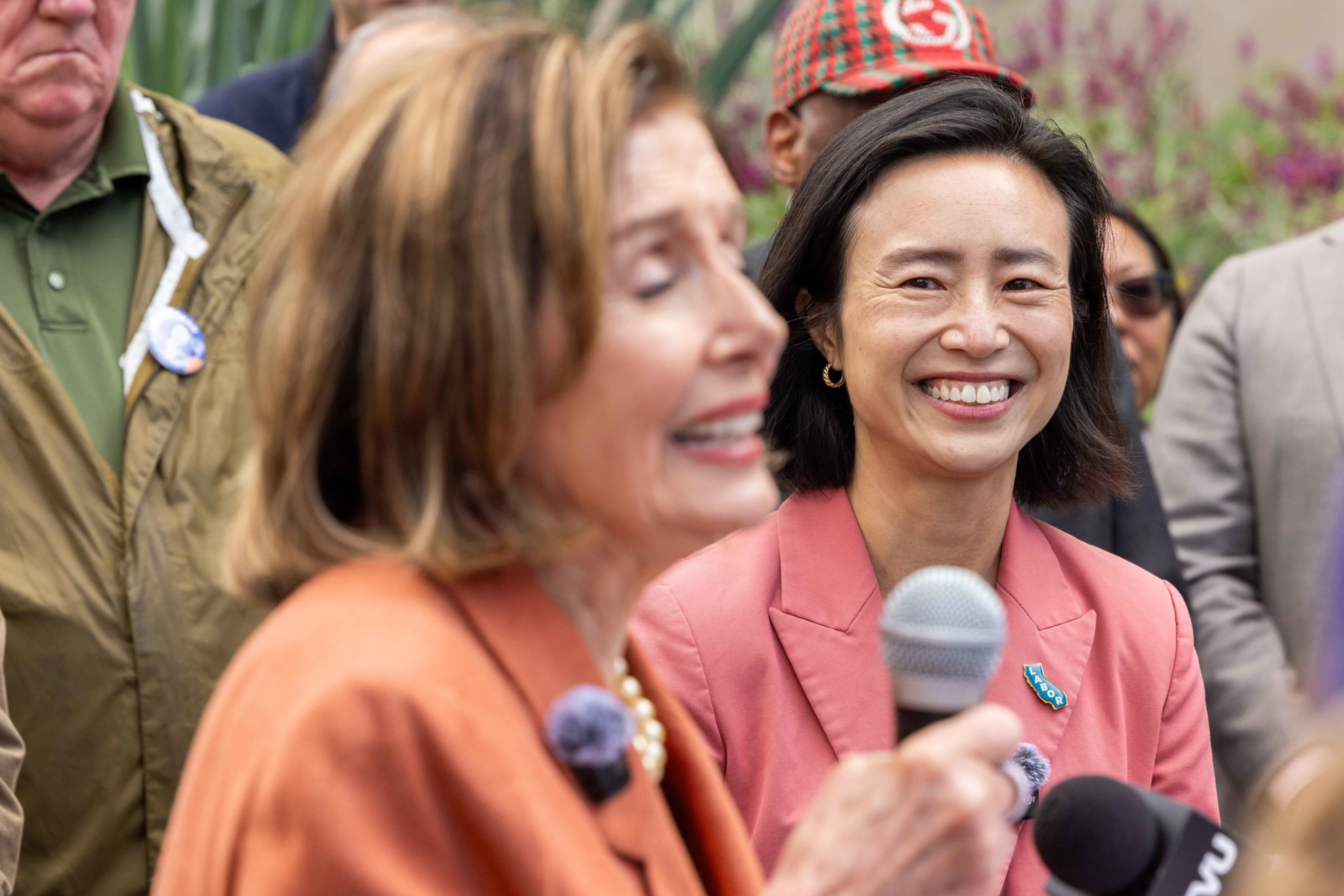 A smiling woman in a pink blazer holds a microphone while another woman in an orange blazer speaks, surrounded by several people in the background.
