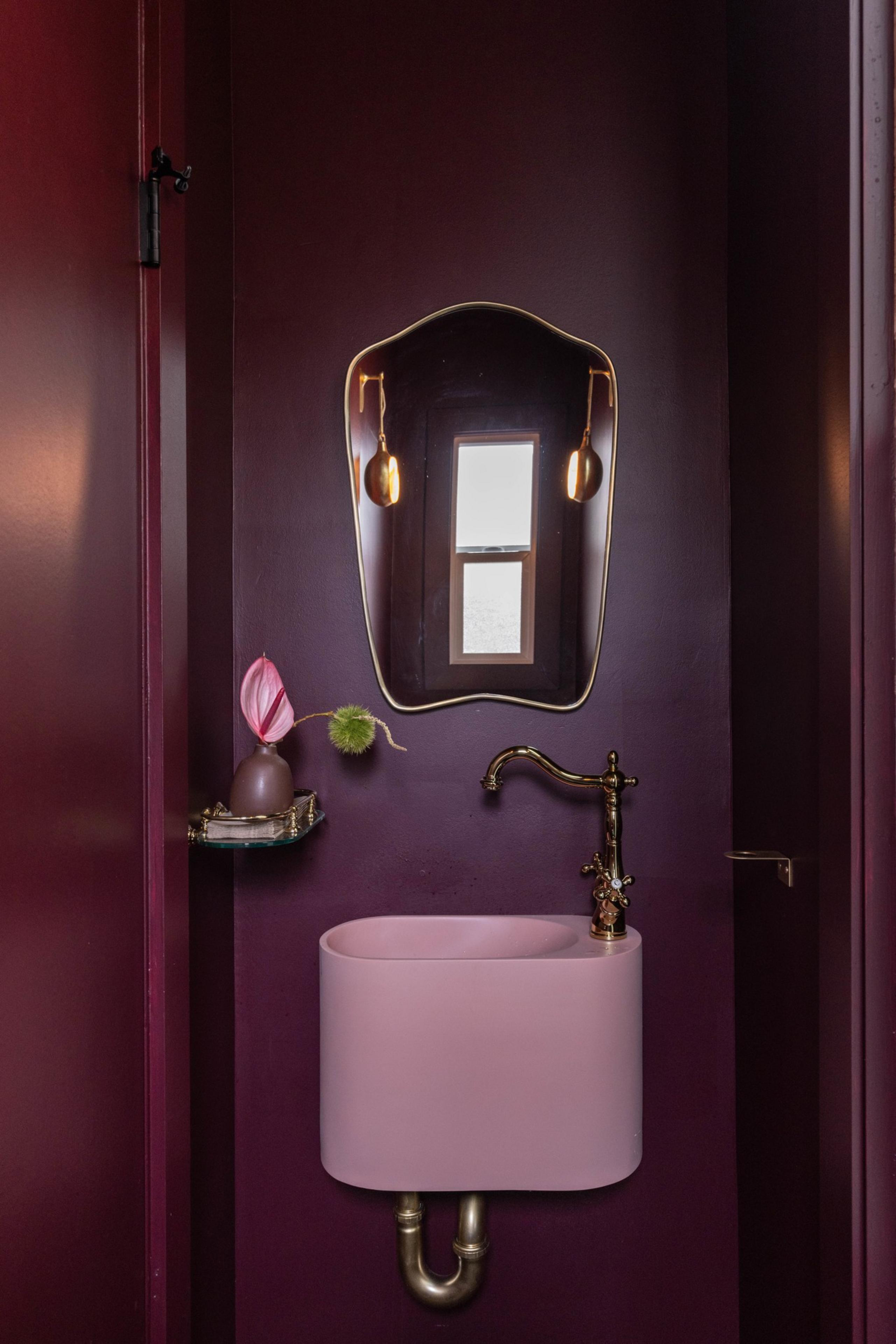 The image shows a small bathroom with dark purple walls, a pink sink, a brass faucet, a decorative mirror, a small shelf with a vase, and a window in the mirror's reflection.