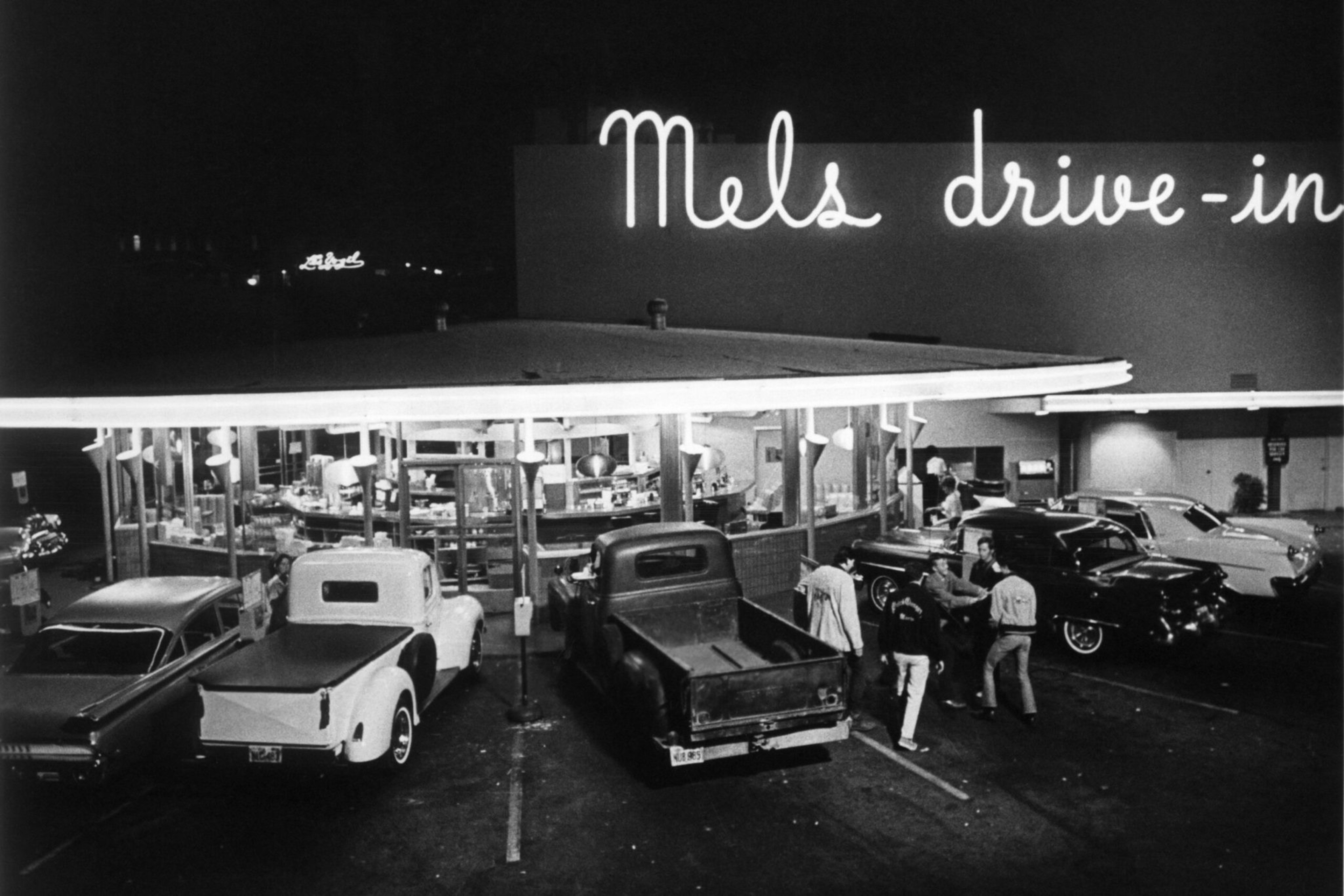 A nighttime scene at Mel's Drive-In shows vintage cars parked with four people standing and talking near a classic black car under bright lights.