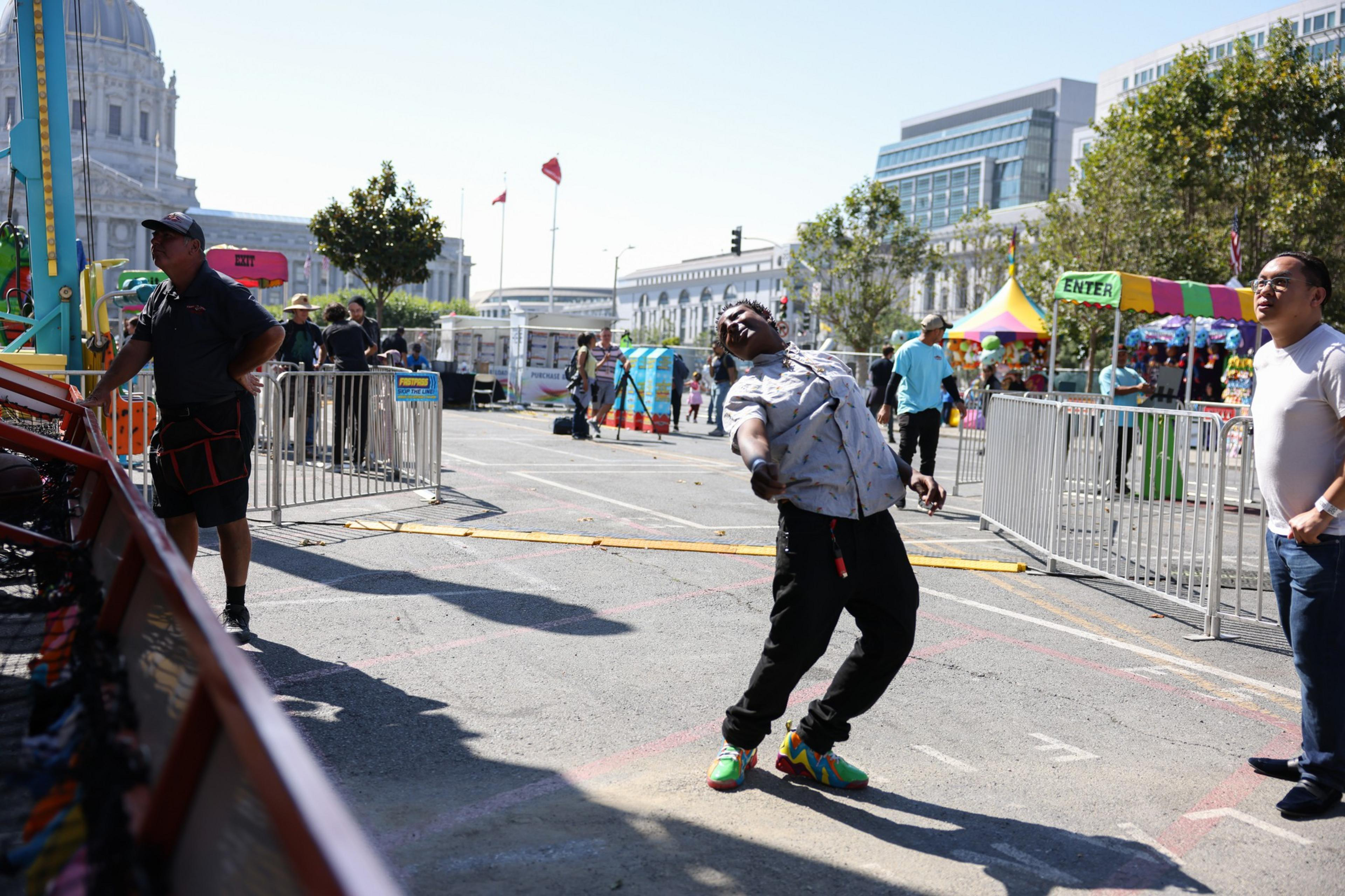 A man leans backward dramatically in an outdoor carnival setting, with colorful shoes, people standing nearby, tents, metal barriers, and buildings in the background.