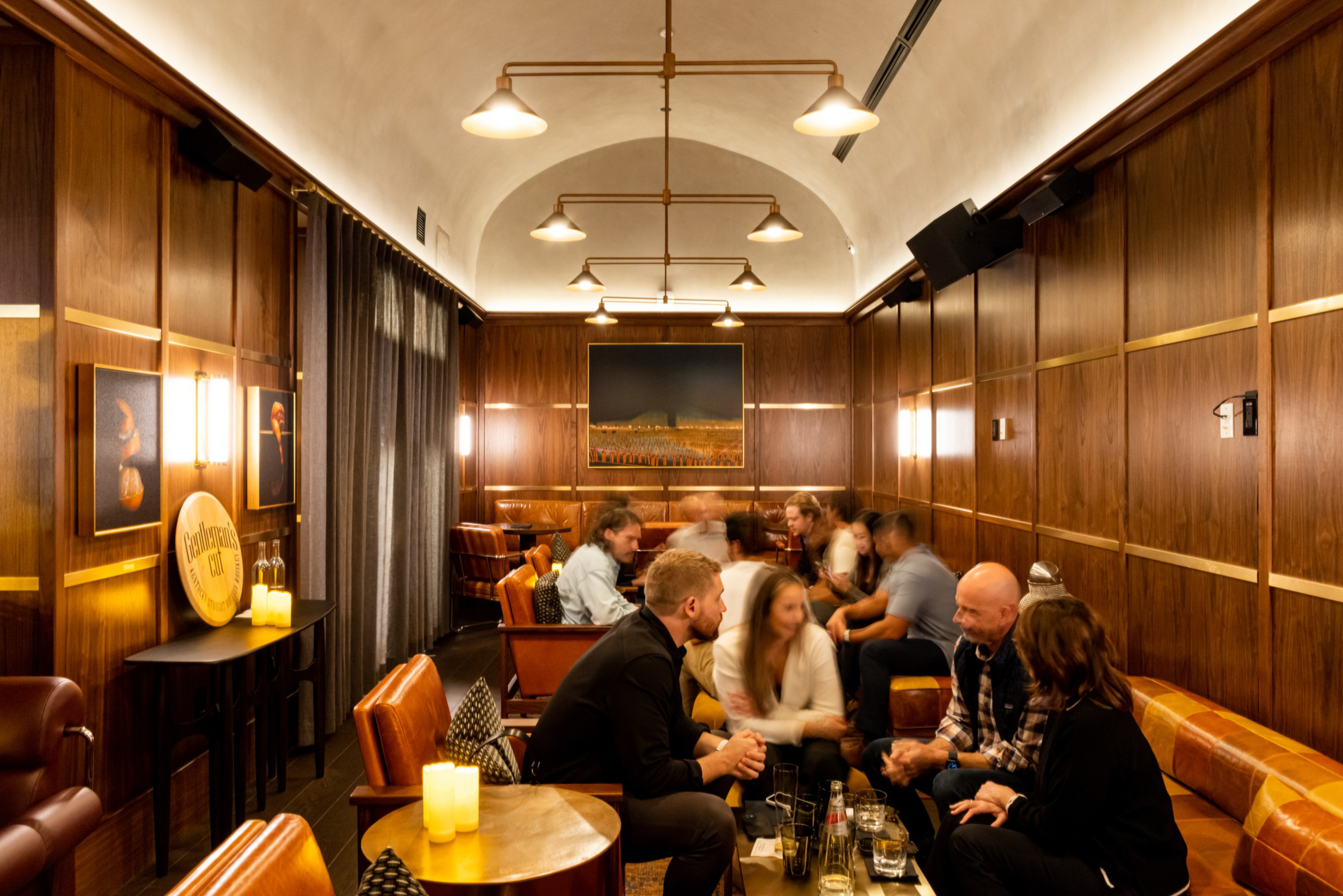 A cozy room with wooden panel walls and warm lighting hosts two groups of people sitting on leather chairs and sofas, engaged in conversation around low tables.