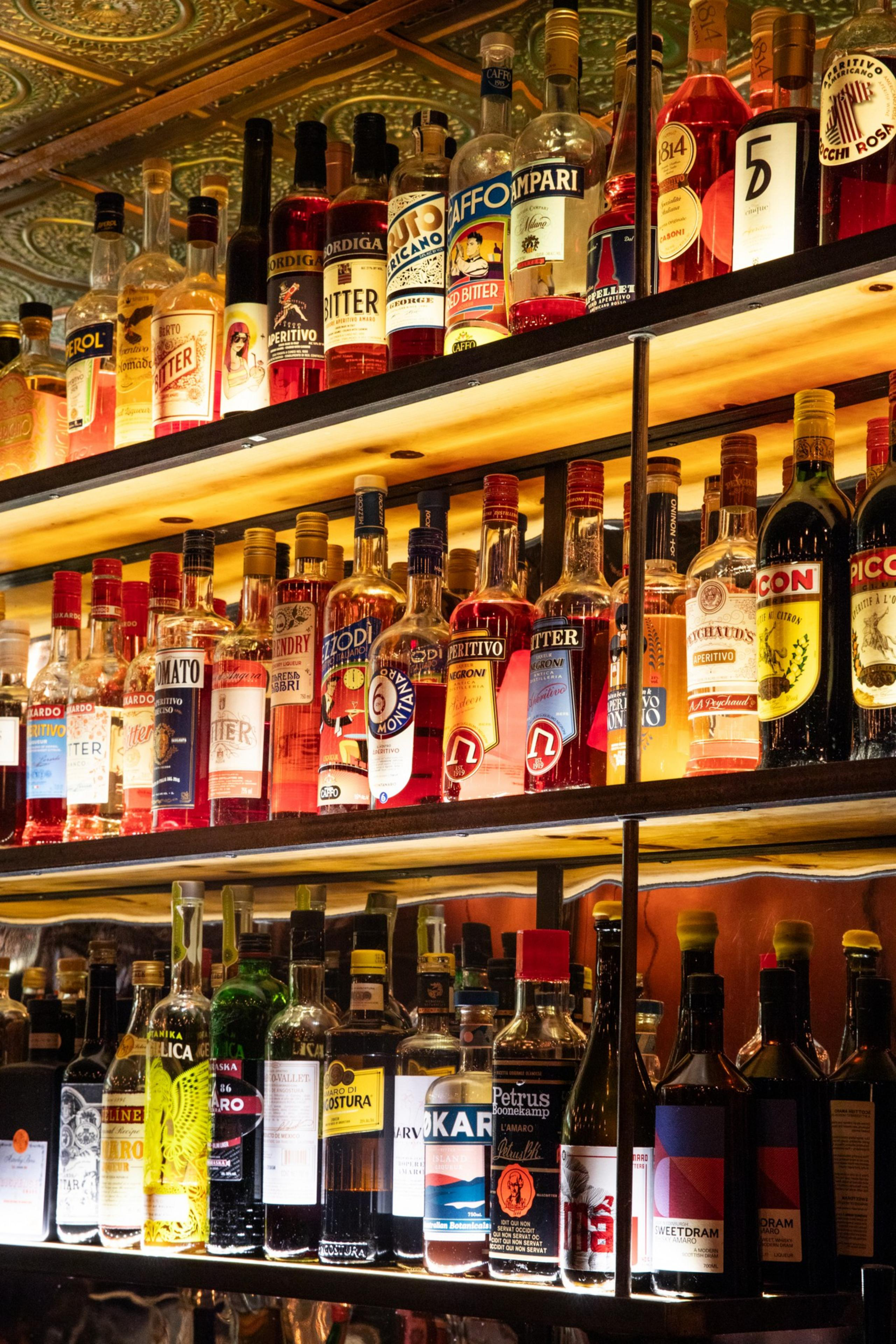 The image shows a backlit bar shelf filled with various brightly colored liquor bottles arranged in rows, featuring diverse labels and designs in vibrant hues.