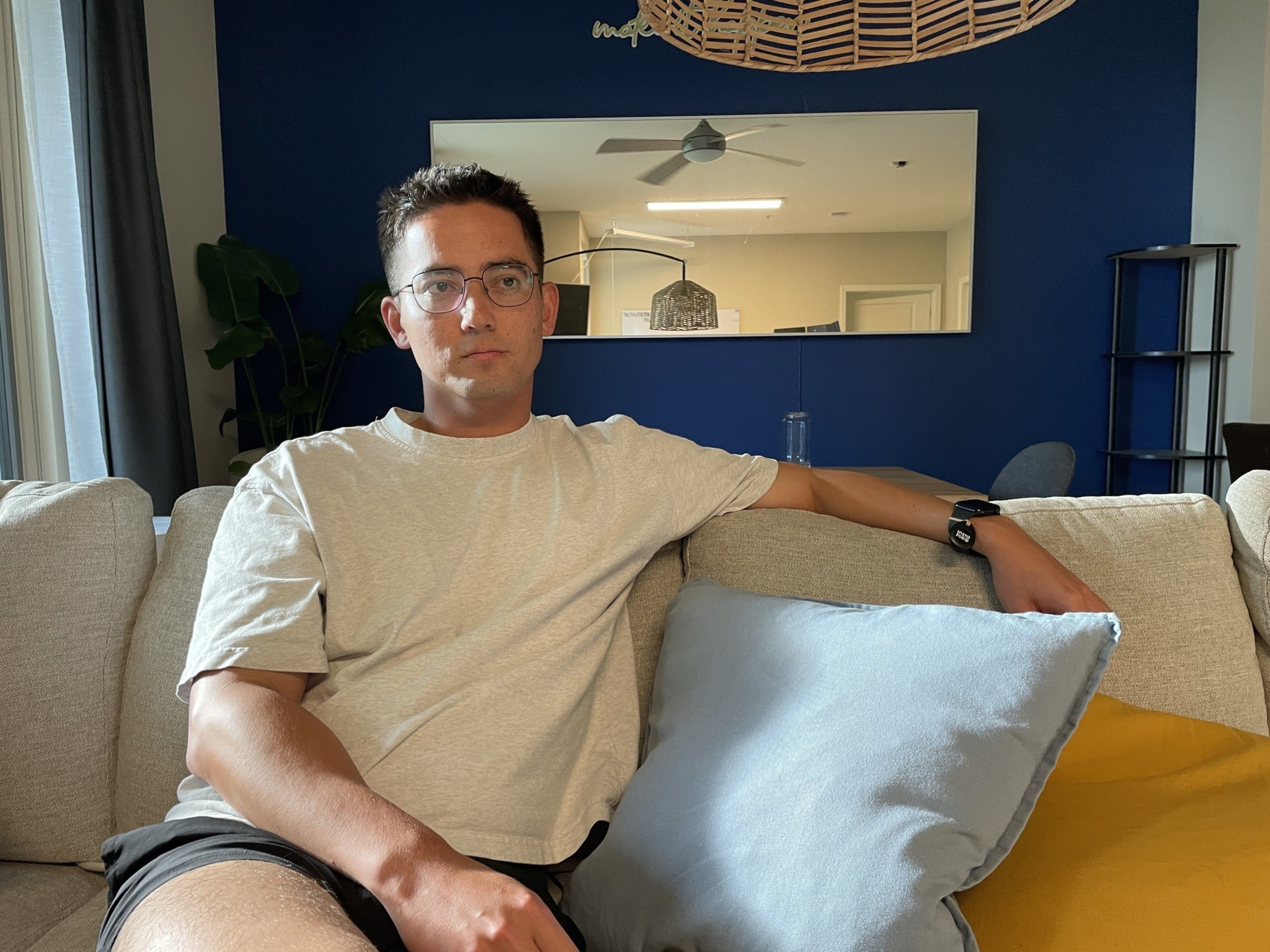 A man wearing glasses and a light beige t-shirt sits on a beige couch with blue and yellow pillows, against a dark blue wall with a mirror and a plant.