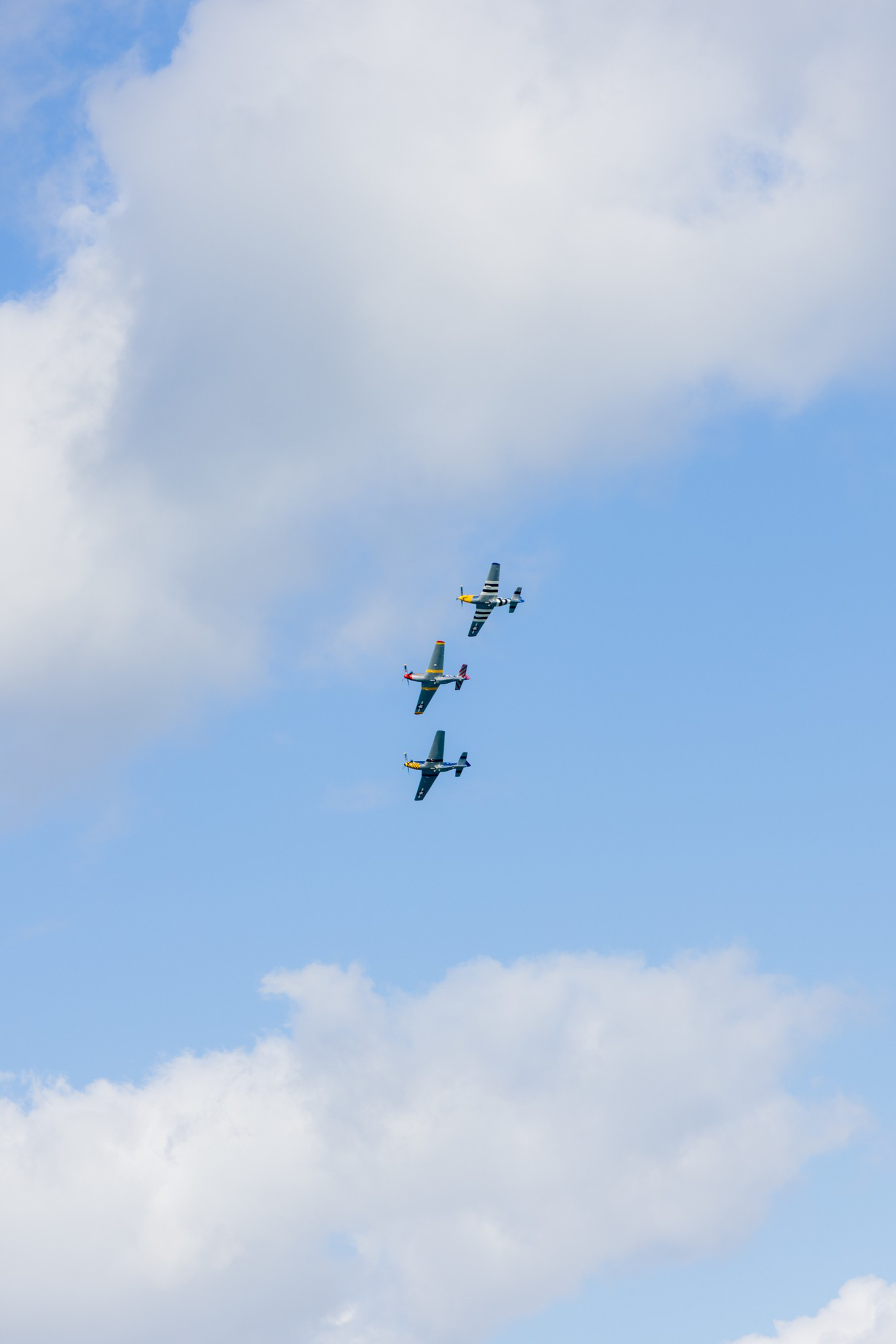 Three vintage military planes fly in close formation against a bright blue sky dotted with large, fluffy white clouds.